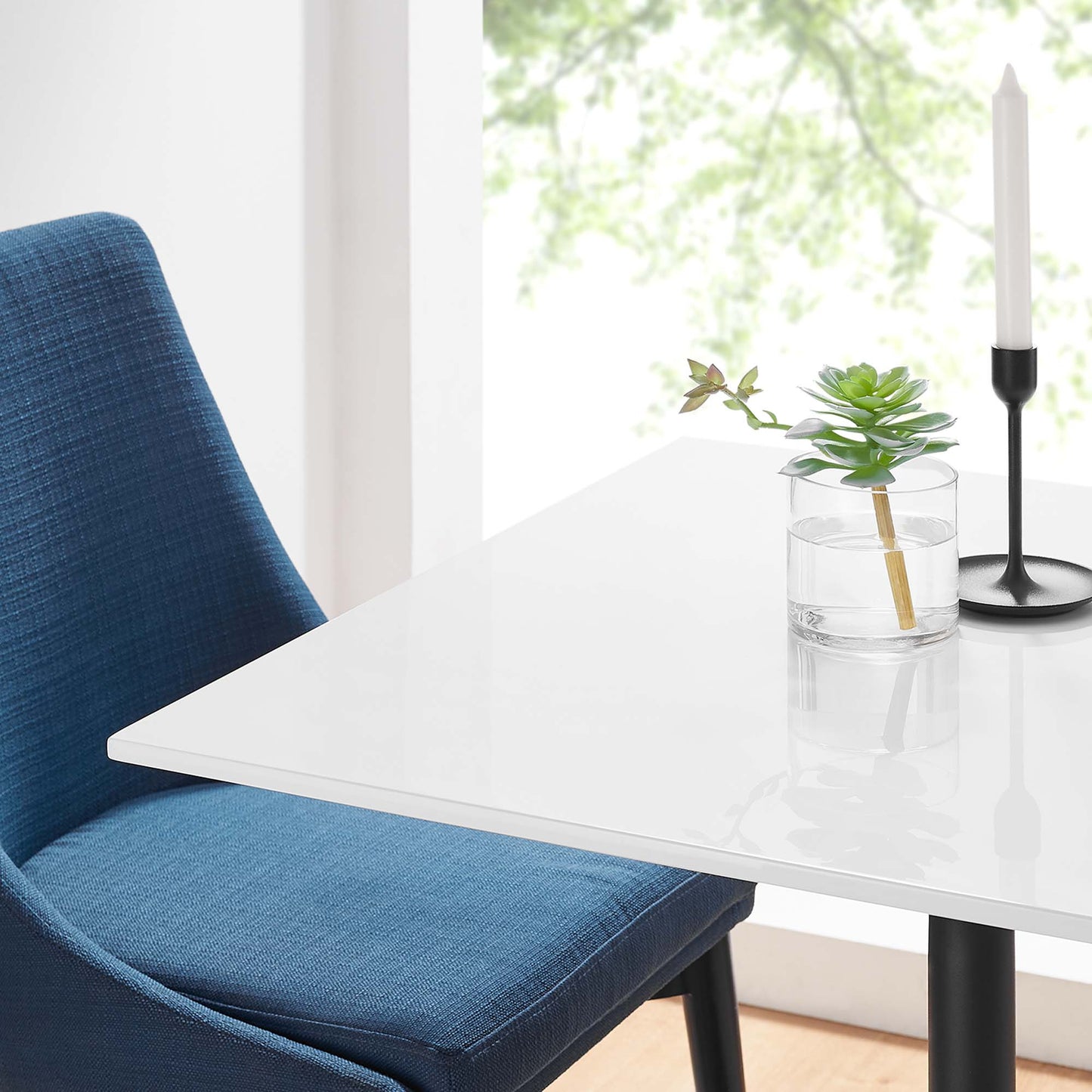 Blue upholstered chair beside a white square wood dining table with a glass vase, succulent, and a black candle holder with a white taper.