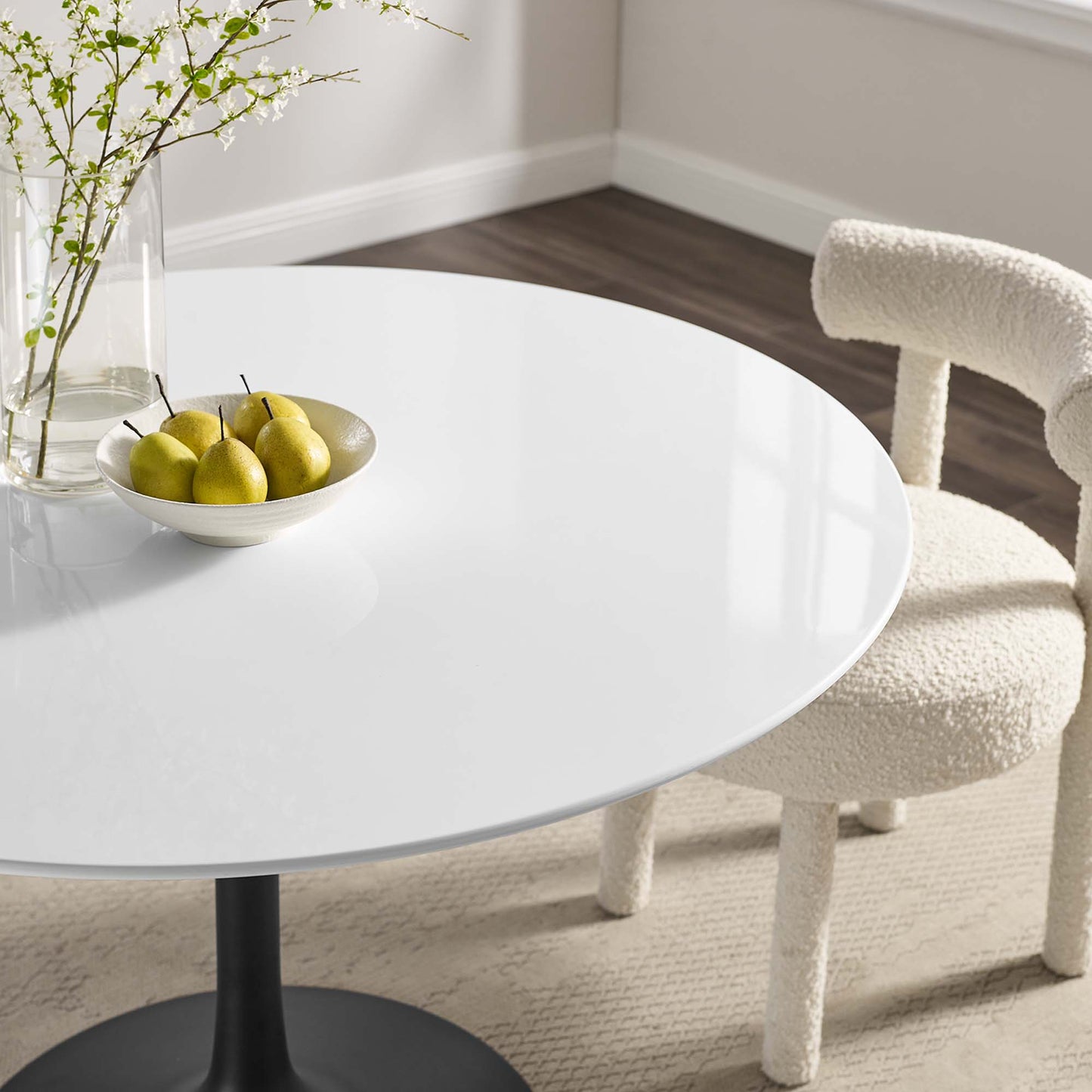 Round wood dining table (48-inch) in a bright room, with a bowl of pears, a glass vase with branches, and a cream boucle chair.