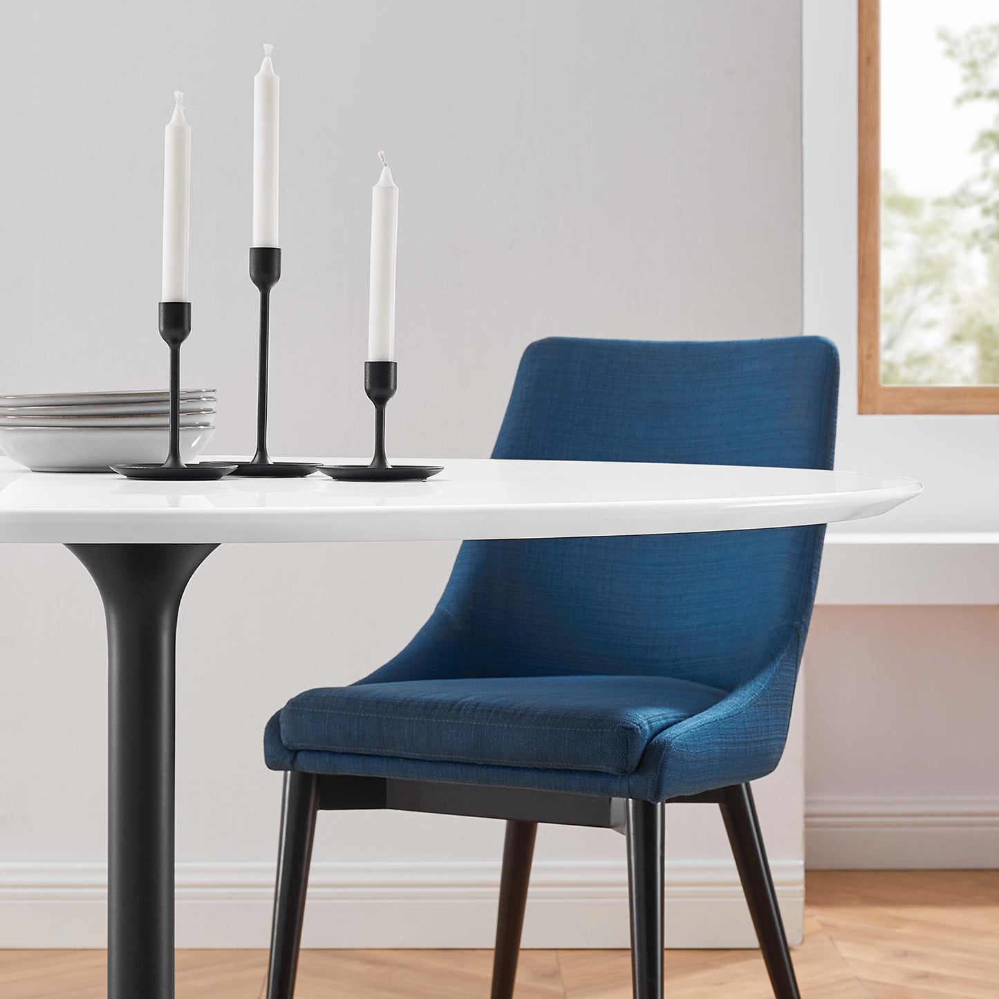 Round white dining table on a black pedestal base, blue upholstered chair, three black candleholders with white candles.
