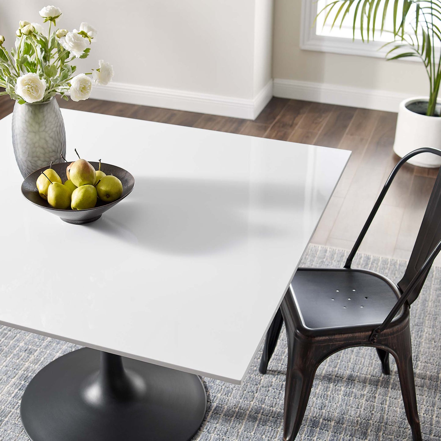 White square wood dining table with black pedestal base, dark metal chair, bowl of pears, and a vase of white flowers.