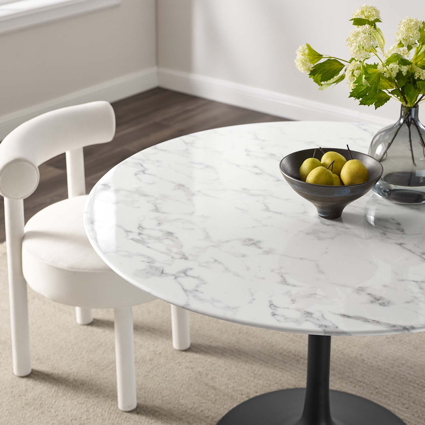 Round faux marble dining table with gray veins on a black pedestal base, white chair nearby, black bowl of lemons & vase of flowers.