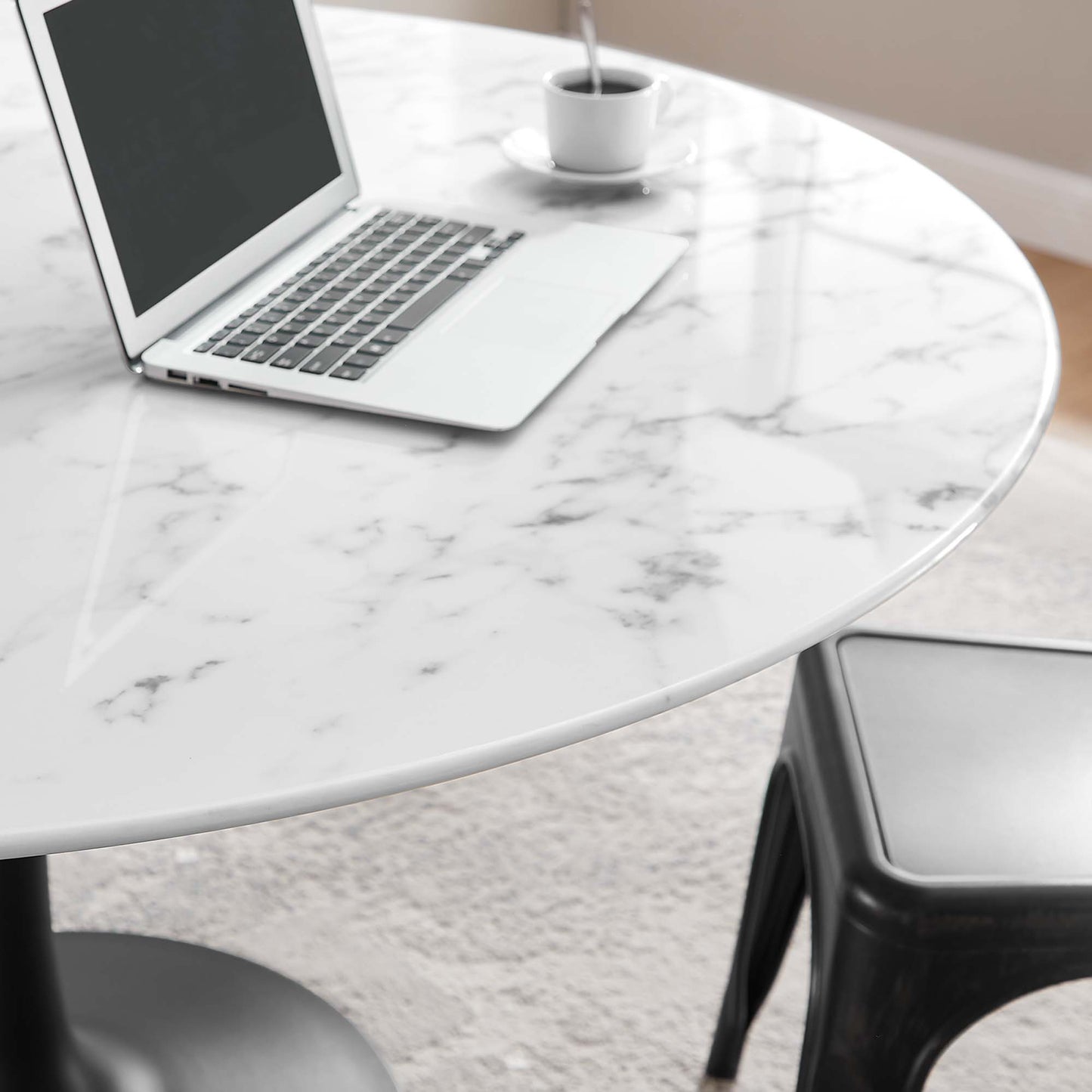 Round faux marble dining table with gray veining, silver laptop, white coffee cup on saucer, black stool beside.