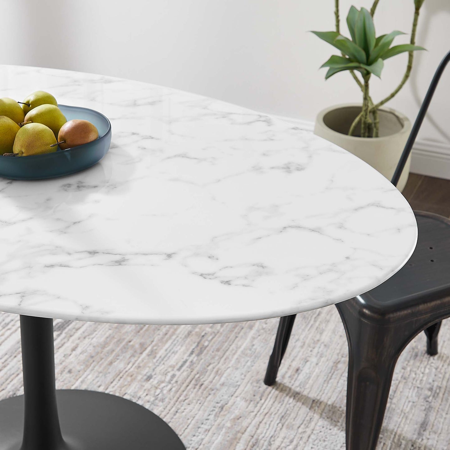 Oval faux marble dining table with black pedestal base; blue fruit bowl with lemons, nearby metal chair and plant.