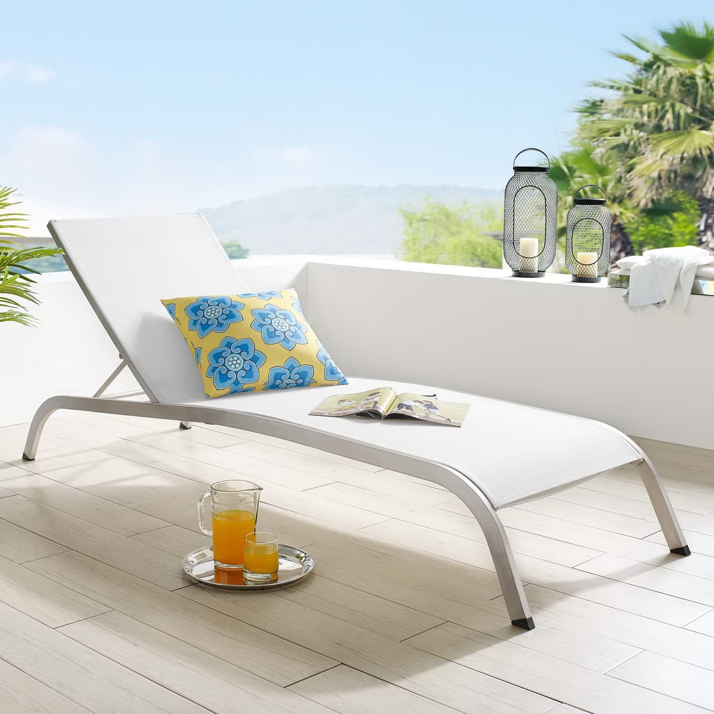 White curved outdoor chaise lounge with blue-yellow floral cushion on sunlit patio; orange juice tray, magazine, lanterns nearby.