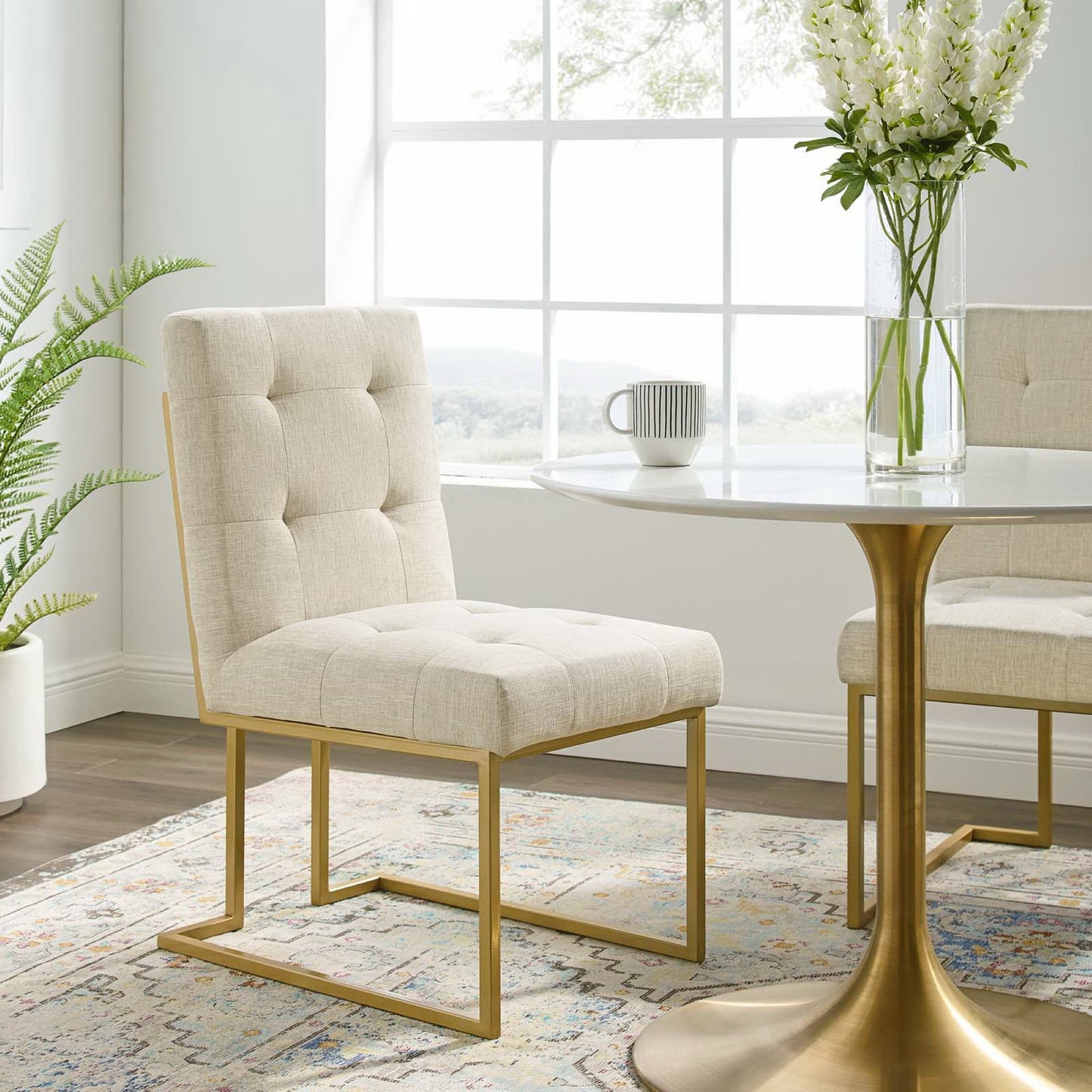Beige tufted dining chair with gold stainless steel legs beside a round white table with a gold pedestal base in a sunlit dining area