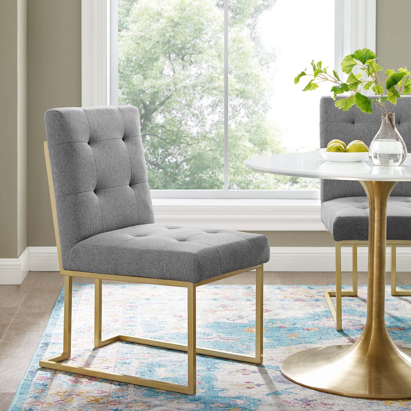 Gray tufted fabric dining chair with a gold stainless steel frame, mid-century style; shown with round white table.