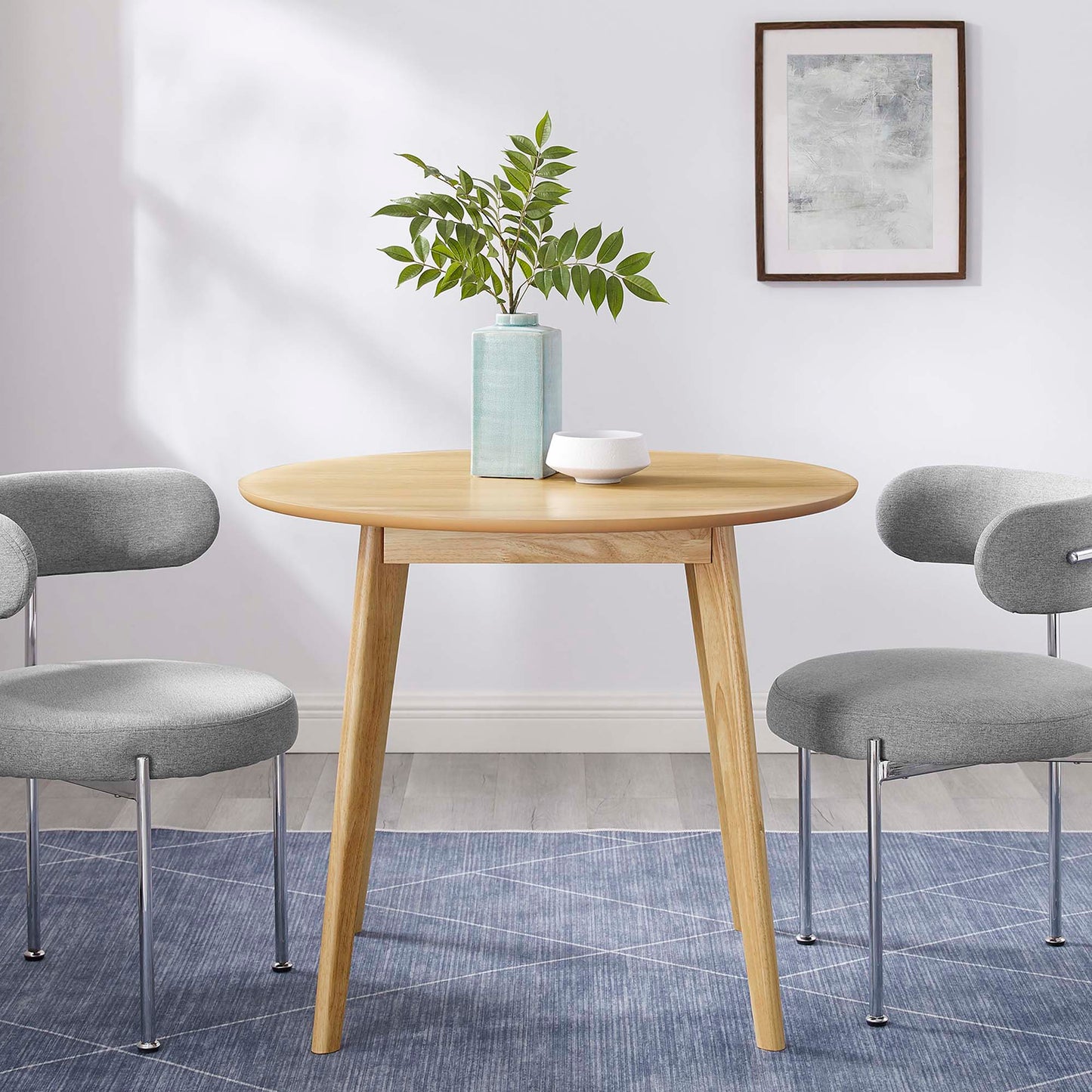 Round dining table with three splayed legs in oak brown finish, gray upholstered chairs, greenery vase and white bowl, mid-century modern style.