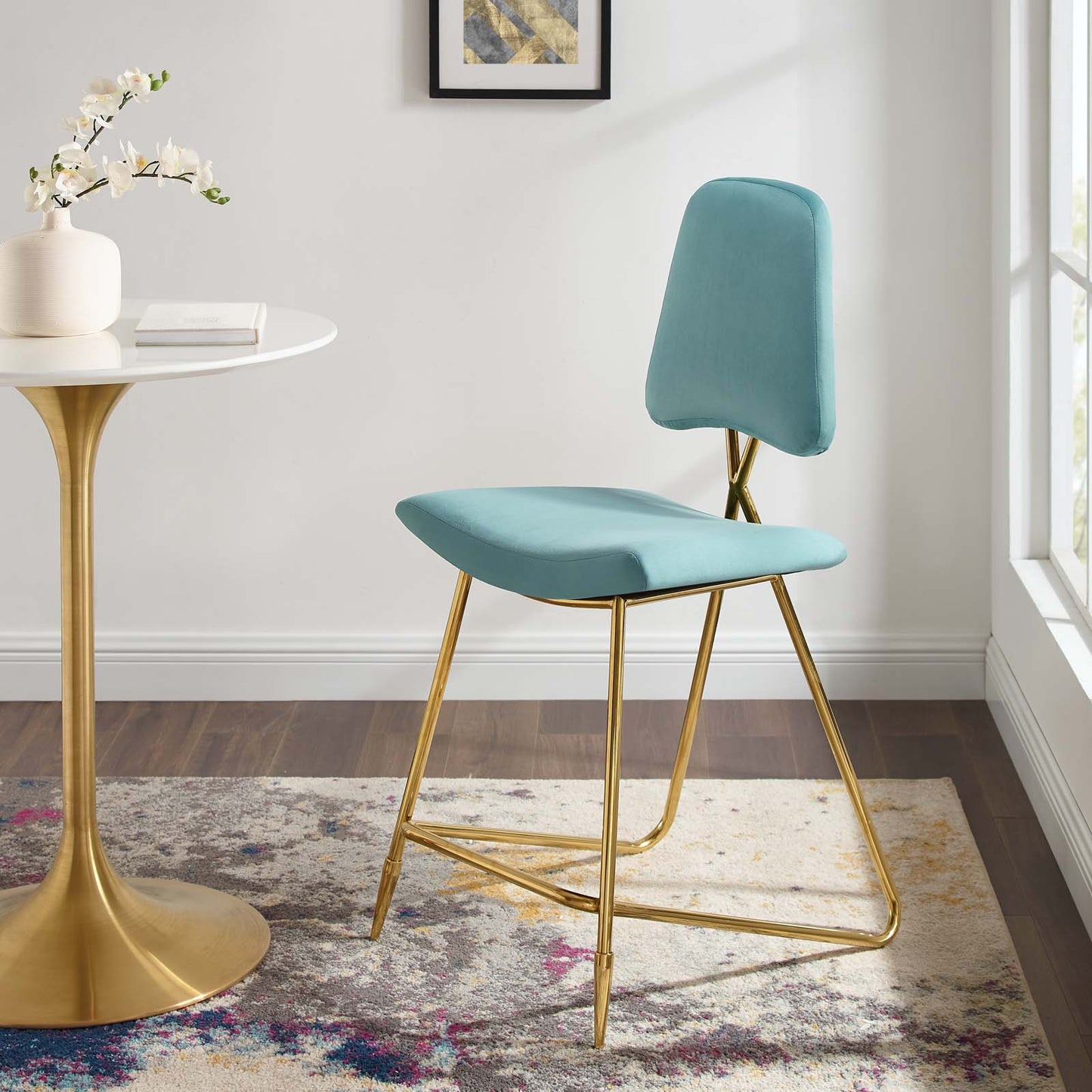 Sea blue velvet counter stool with gold metal legs beside a round white table with a gold pedestal base in a room.