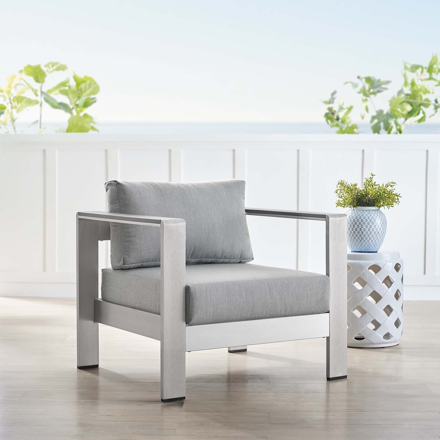 Modern outdoor armchair with a silver-gray aluminum frame and light gray cushions on a sunlit deck, with a lattice side table and a potted plant.