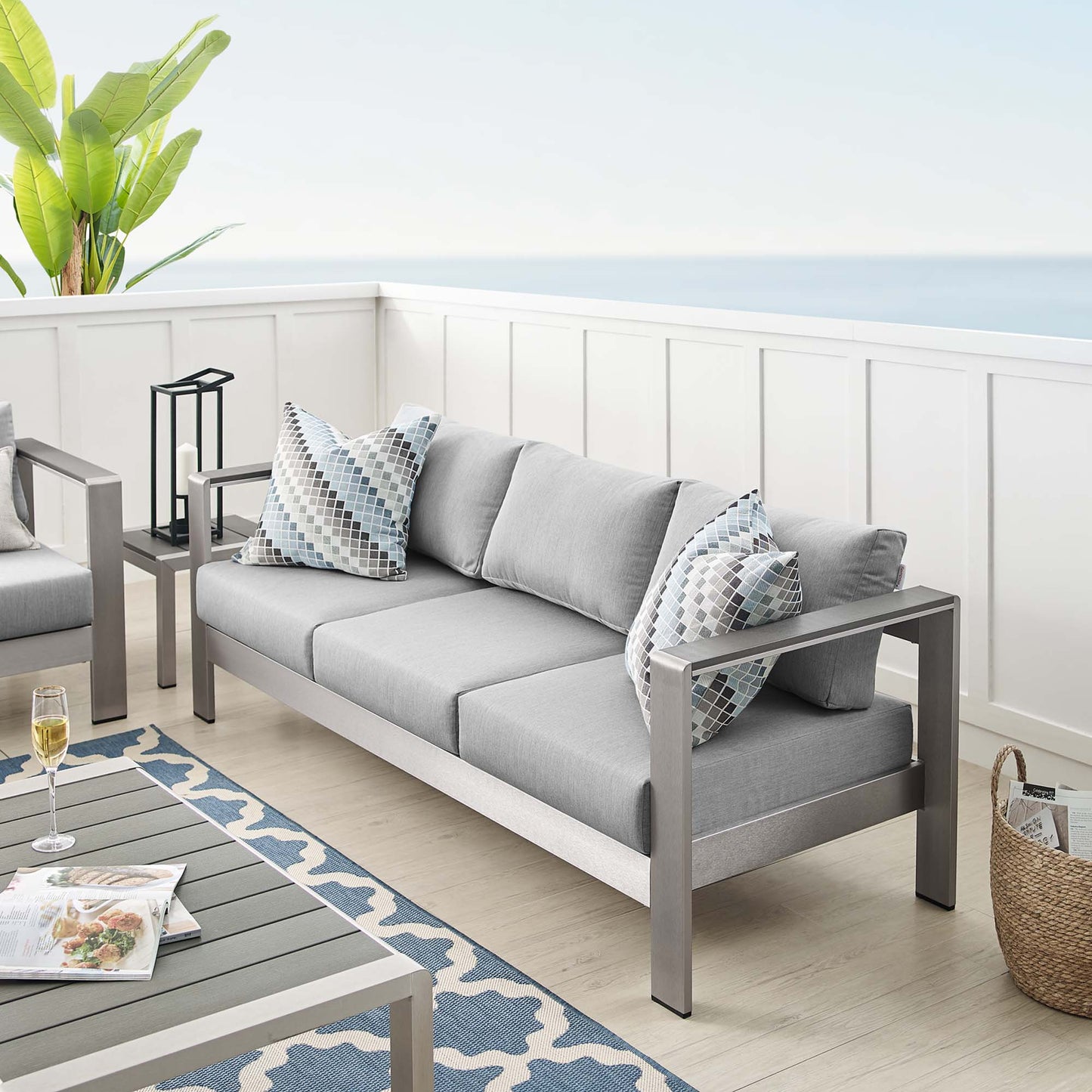 Slate gray outdoor sofa with light gray cushions and geometric-pattern pillows on a modern metal-frame patio set by the sea.