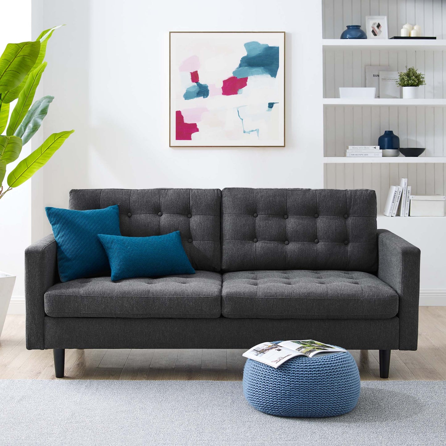 Charcoal gray tufted fabric sofa with two teal cushions, wooden legs, a blue knitted pouf, abstract art, and white shelving.
