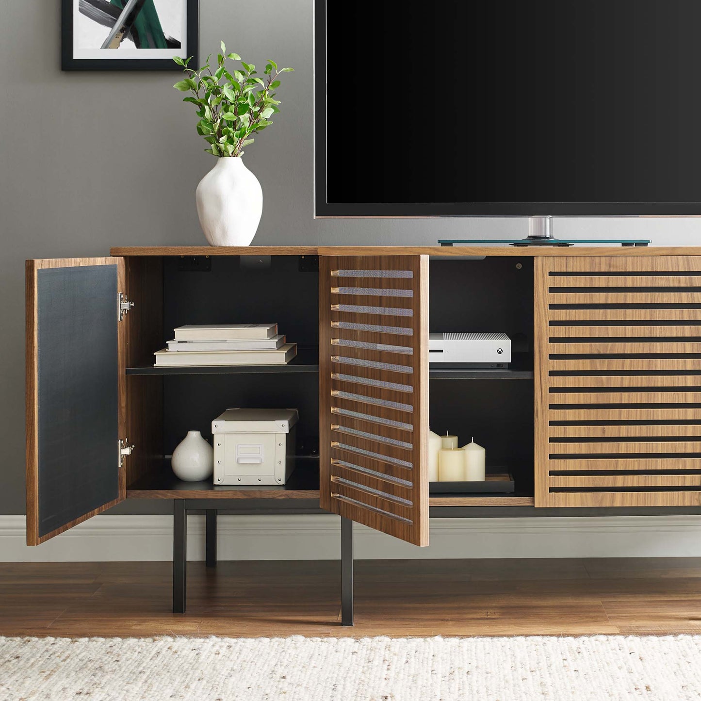 Walnut wood sideboard with black interior, slatted doors, open shelves, TV on top, white vase and greenery.