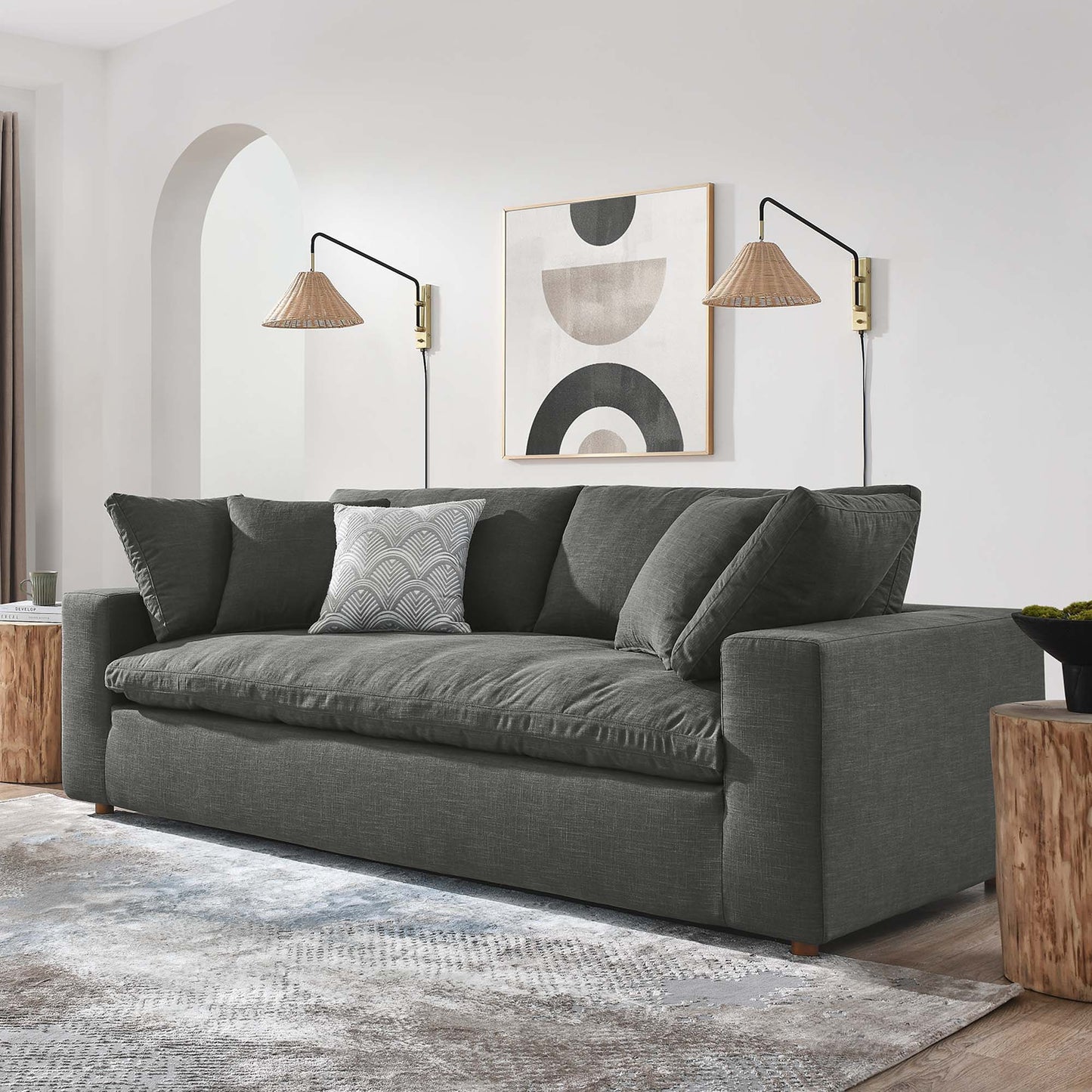 Gray fabric three-seater sofa with plush cushions in a modern living room, flanked by wooden stump side tables and abstract art.
