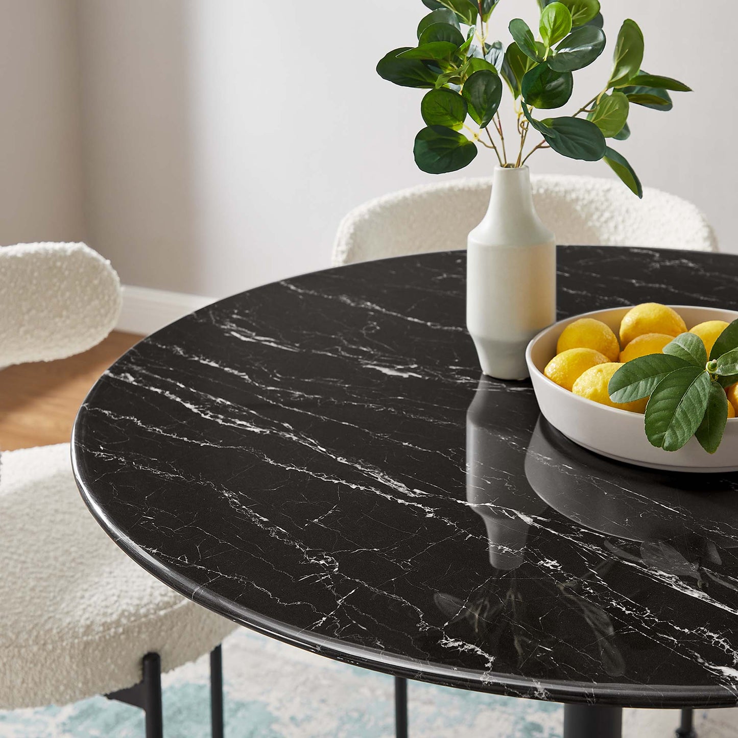 Round black marble table with white veins, white vase with greenery, bowl of lemons in modern dining setting.