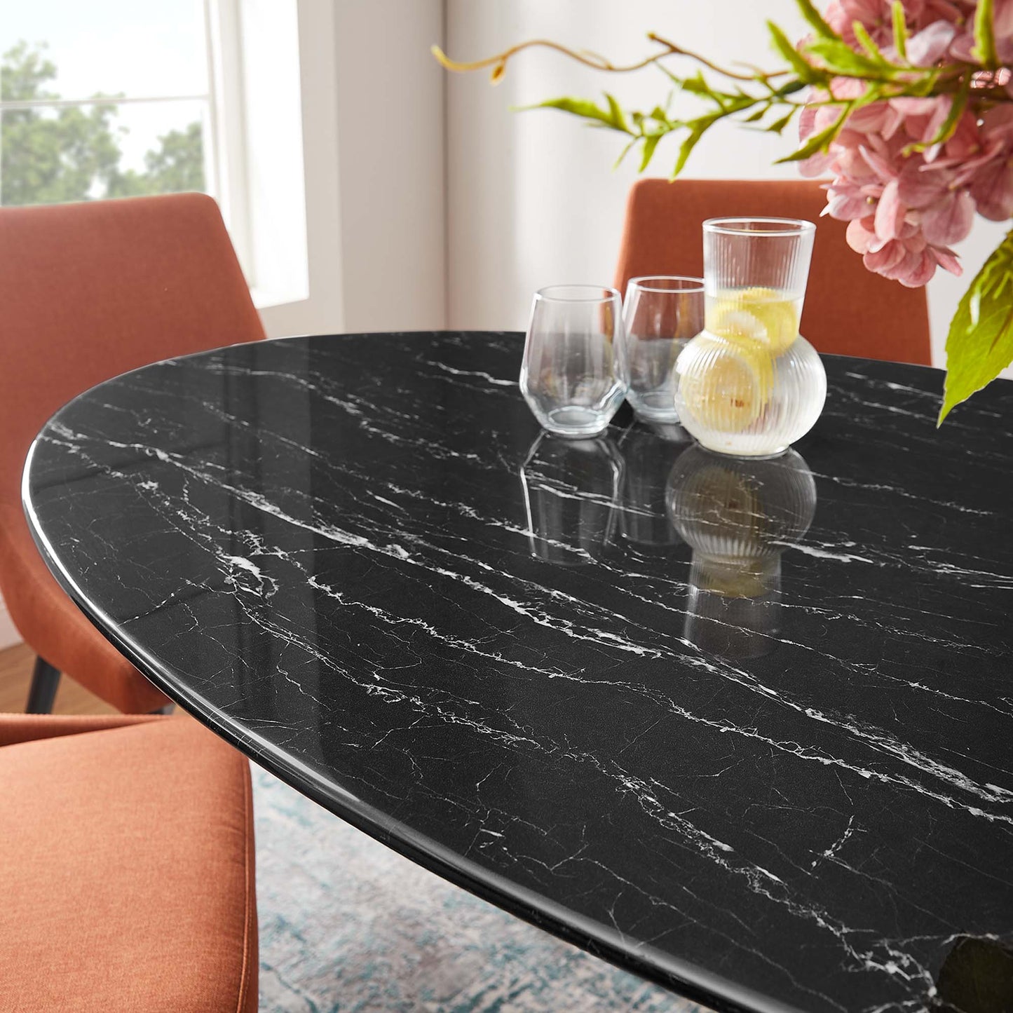 Oval black faux-marble dining table with white veining, orange upholstered chairs, glass tumblers, a glass vase with lemons, pink flowers.