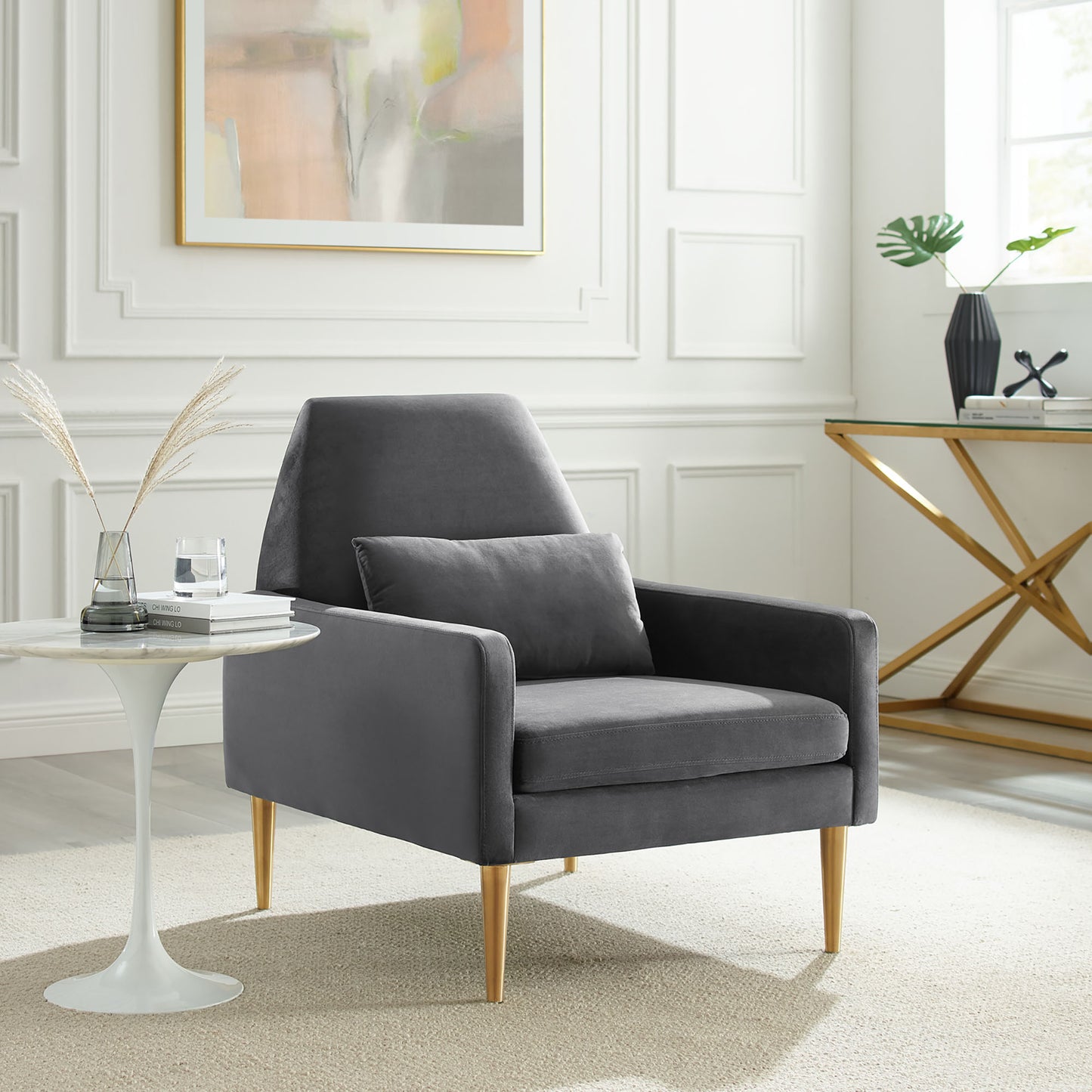 Grey velvet armchair with gold tapered legs and matching cushion in a bright paneled living room.