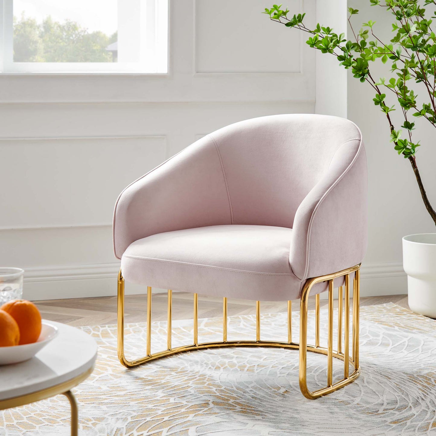 Blush pink velvet accent chair with gold curved metal legs and soft rounded arms in a bright living room.