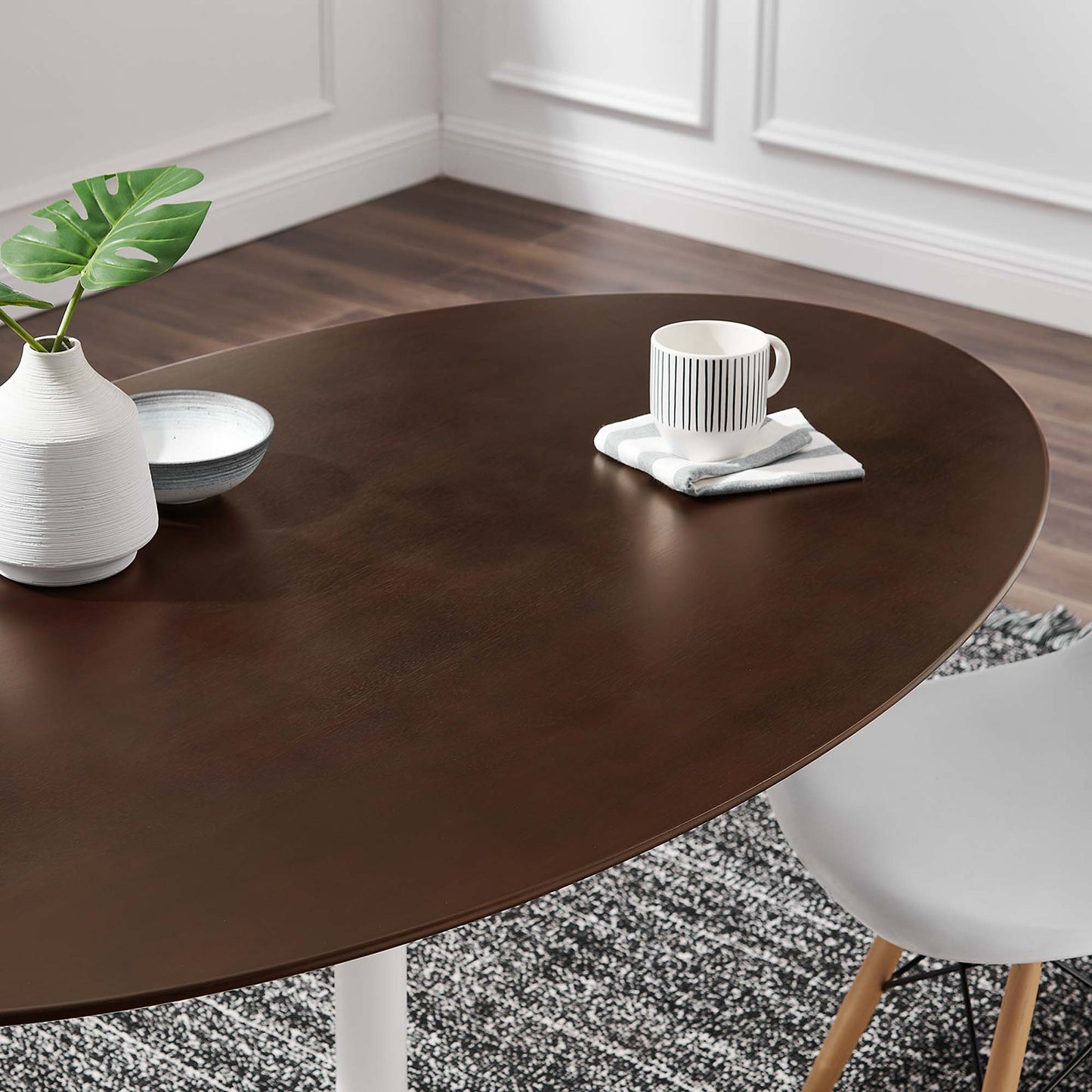 Oval dark brown wood dining table with white legs, white chair with wooden legs, mug on napkin, plant vase and bowl.