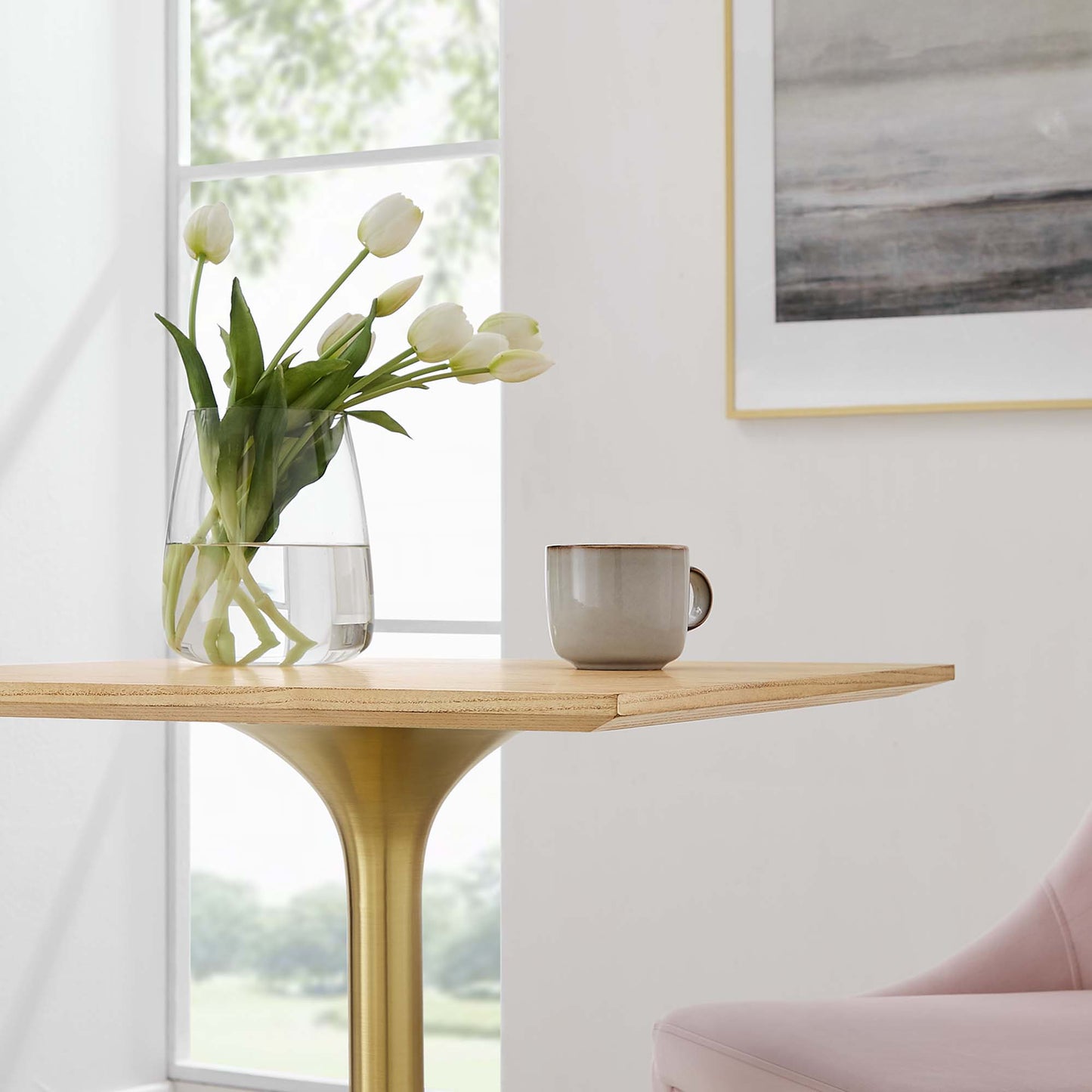 Gold pedestal table with natural wood top, glass vase of white tulips and a mug in a bright room.