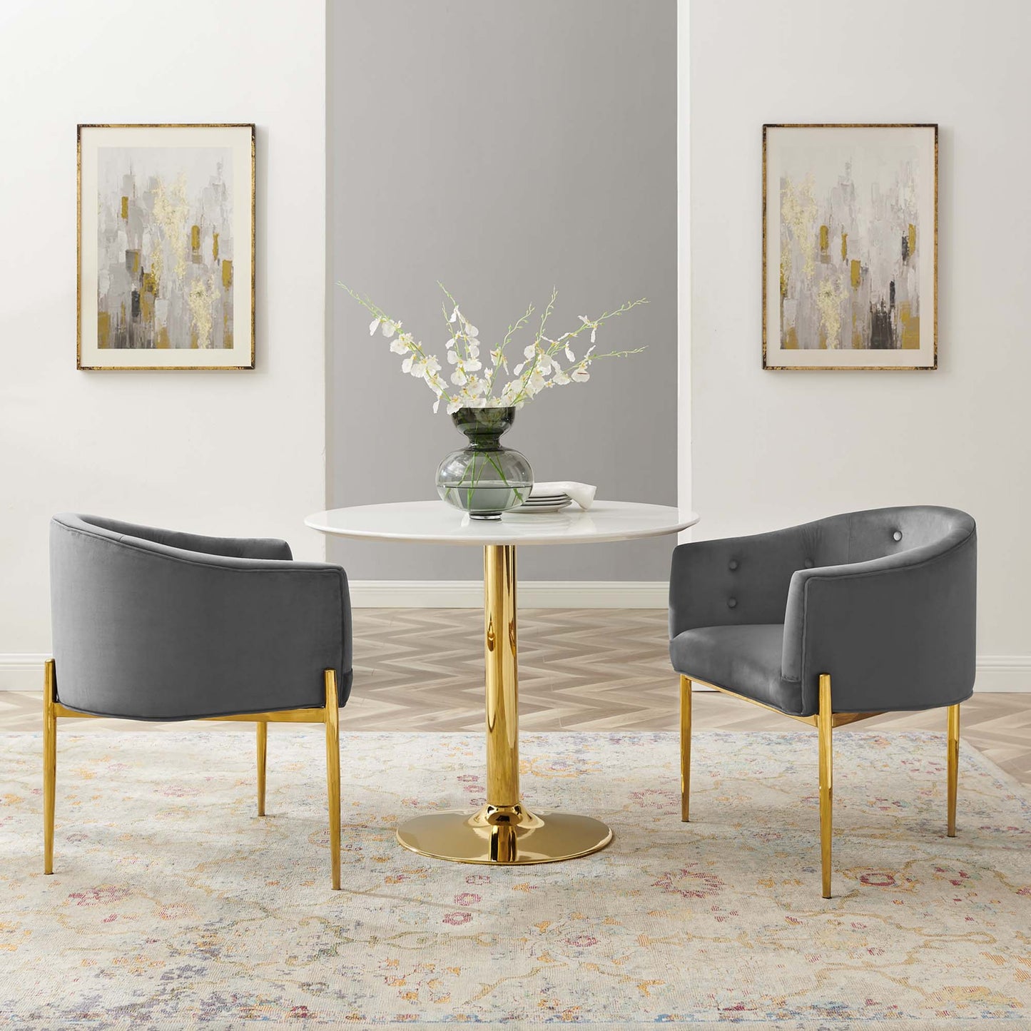 Round white table with gold pedestal, two gray curved chairs with gold legs, glass vase of white flowers, abstract wall art.