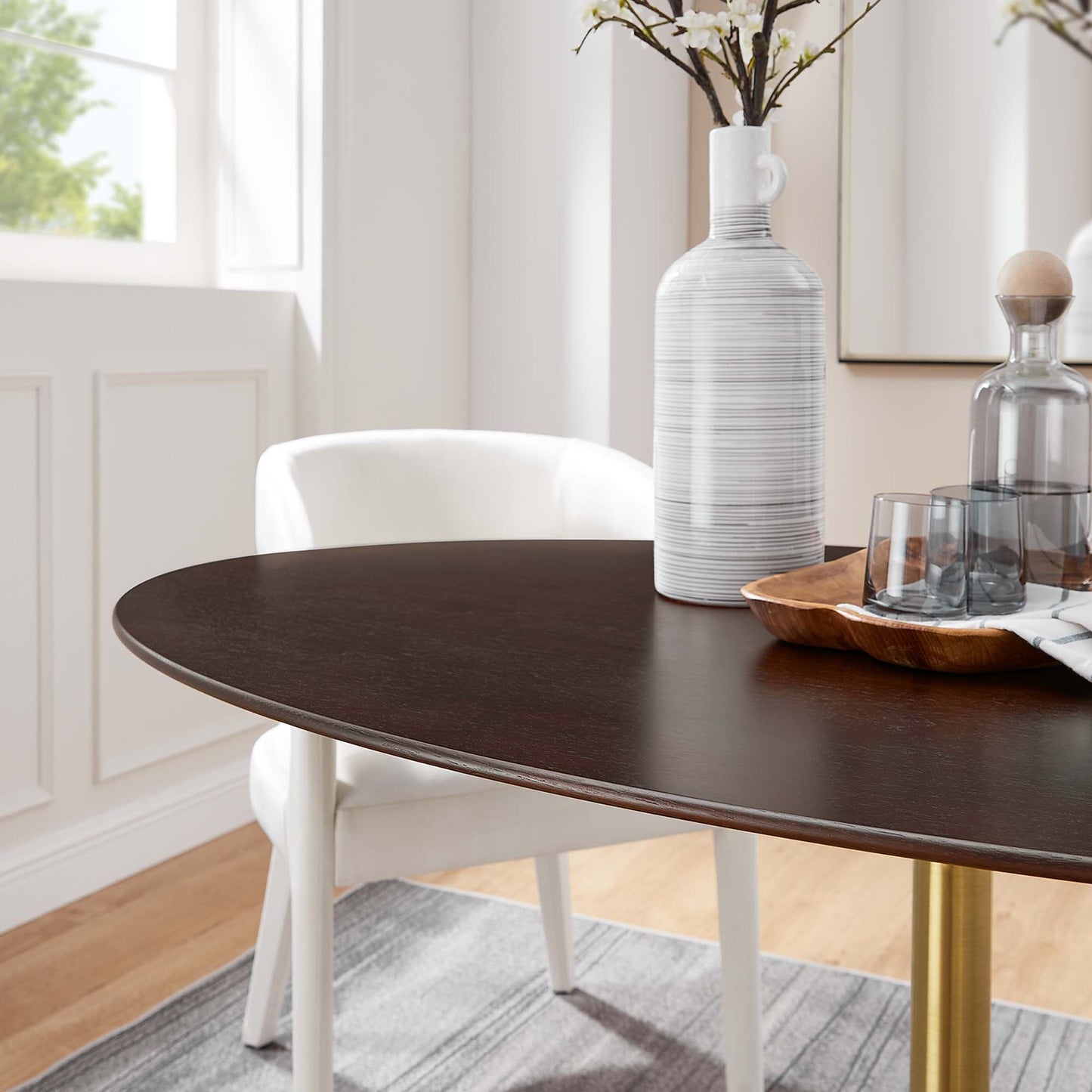Oval dark wood dining table with a gold leg, white ribbed ceramic vase, wooden tray and glassware in a bright modern dining room.
