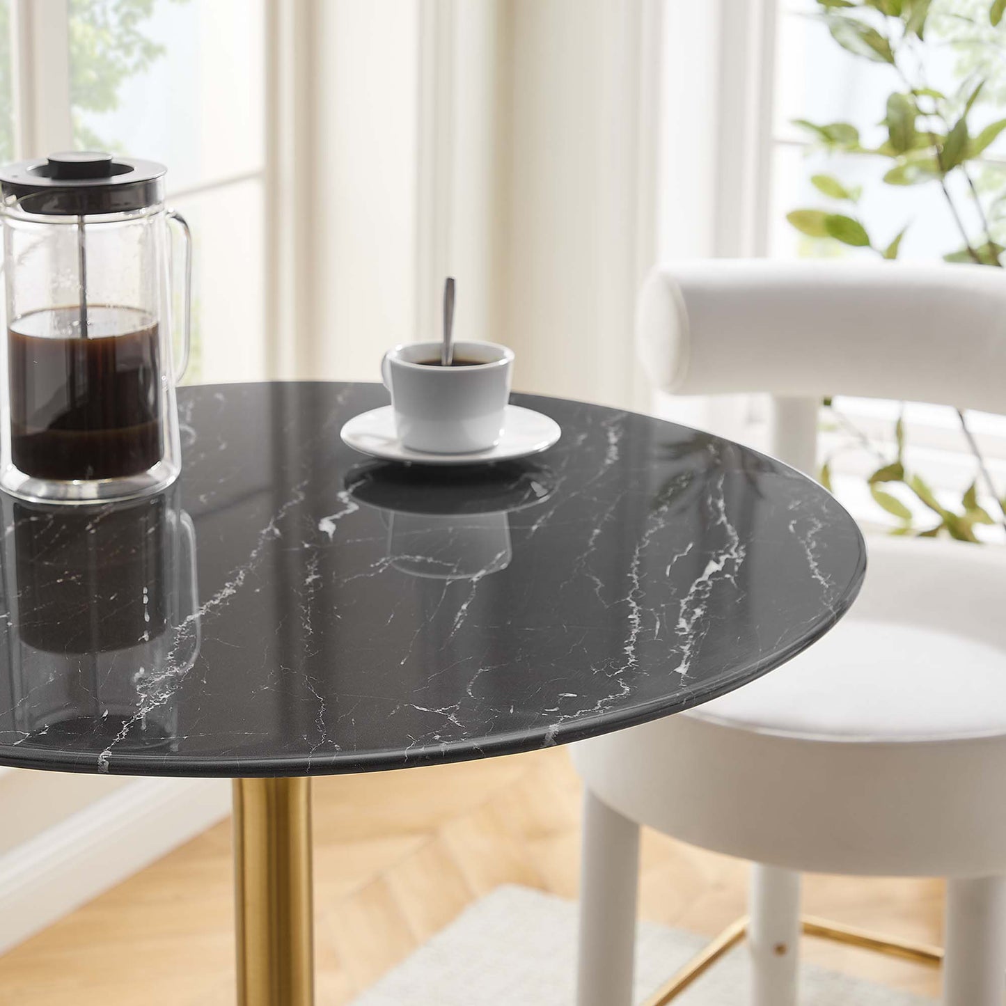 Round black faux marble bar table with a gold pedestal; glass coffee pot and white espresso cup on saucer in a bright modern dining area.