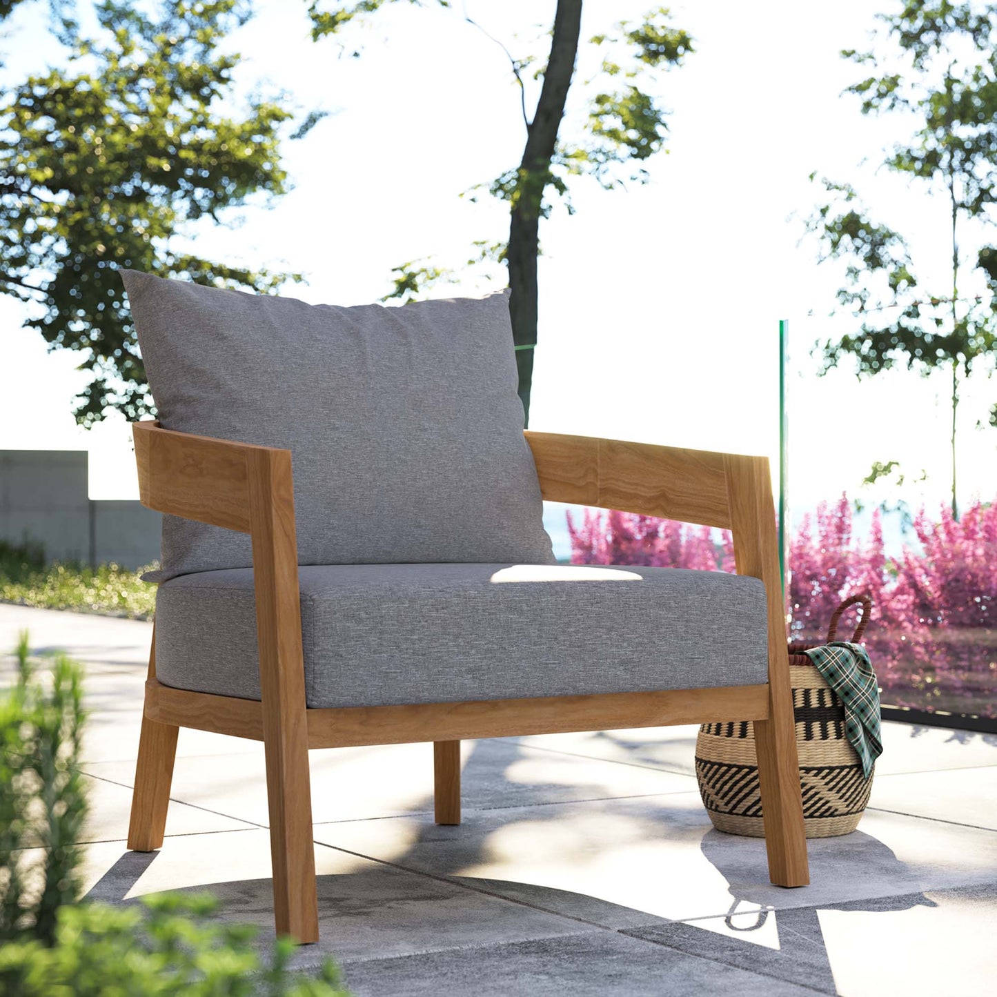 Outdoor teak wood patio armchair with light gray cushions on a sunlit patio, with a woven basket beside.
