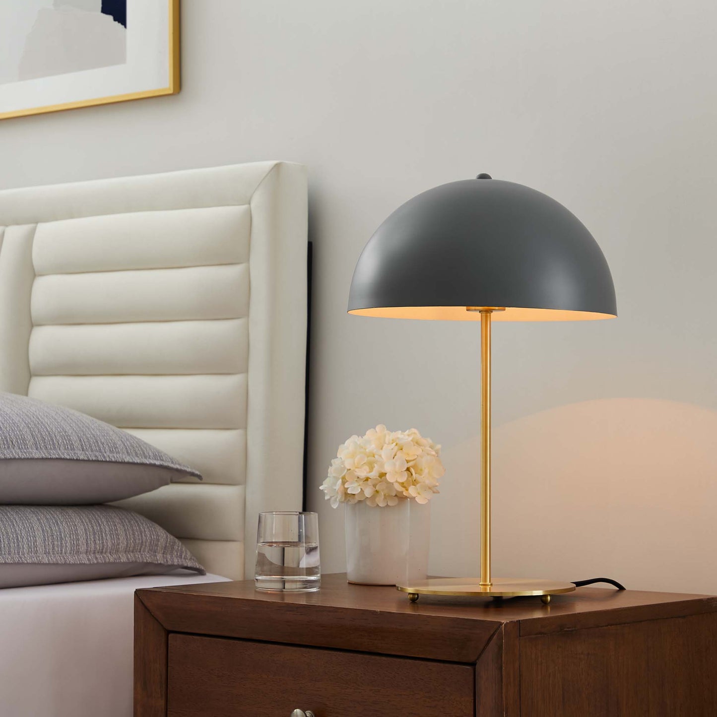 Brown dome lamp with a gold stem on a dark wood nightstand beside a white tufted headboard, with a glass and a vase of cream flowers