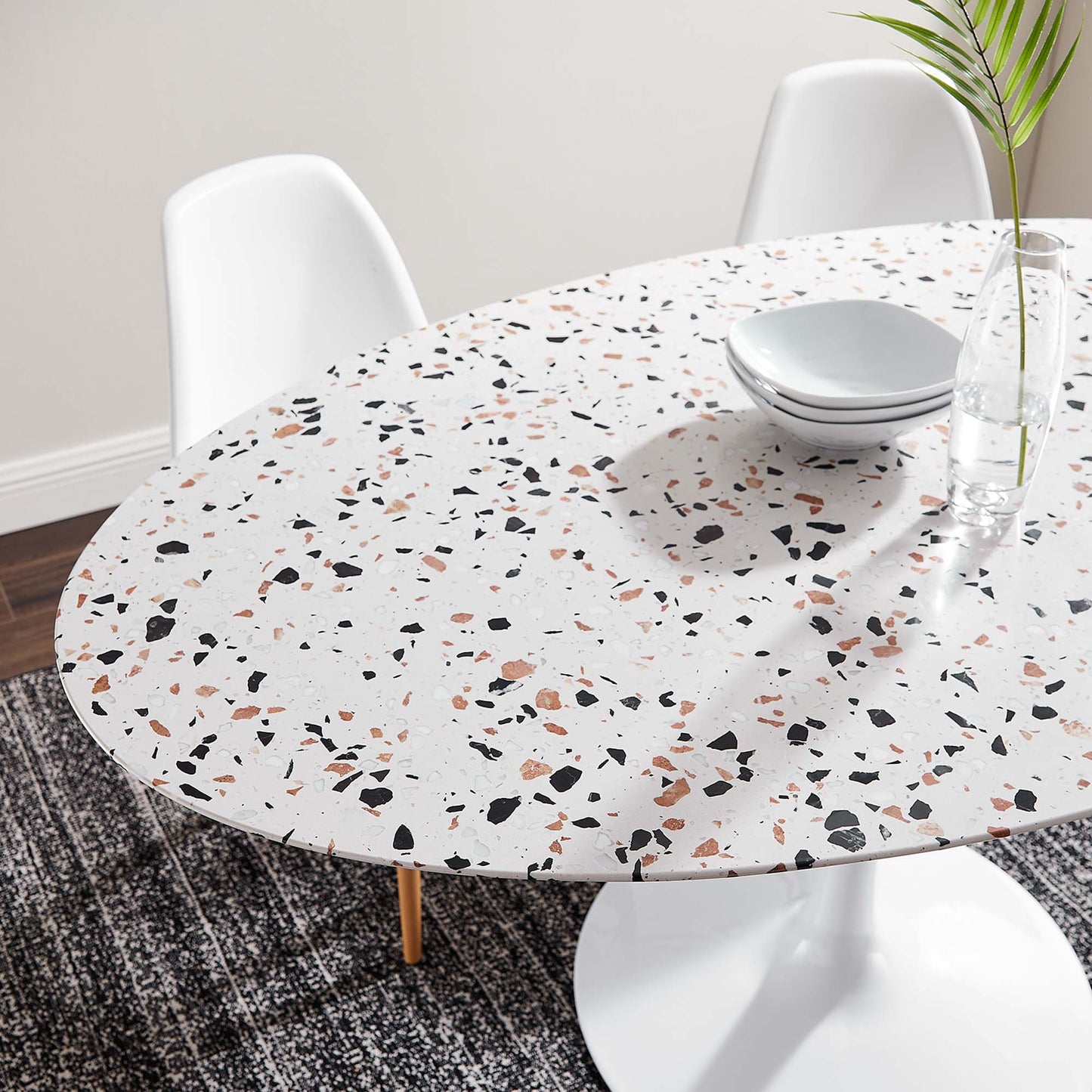 Oval terrazzo-white dining table with multicolor speckles, white chairs, plates and glass vase with plant.