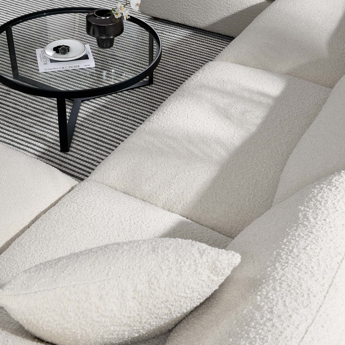 Ivory boucle sectional sofa with matching pillow, round glass-top black metal coffee table on striped grey rug.