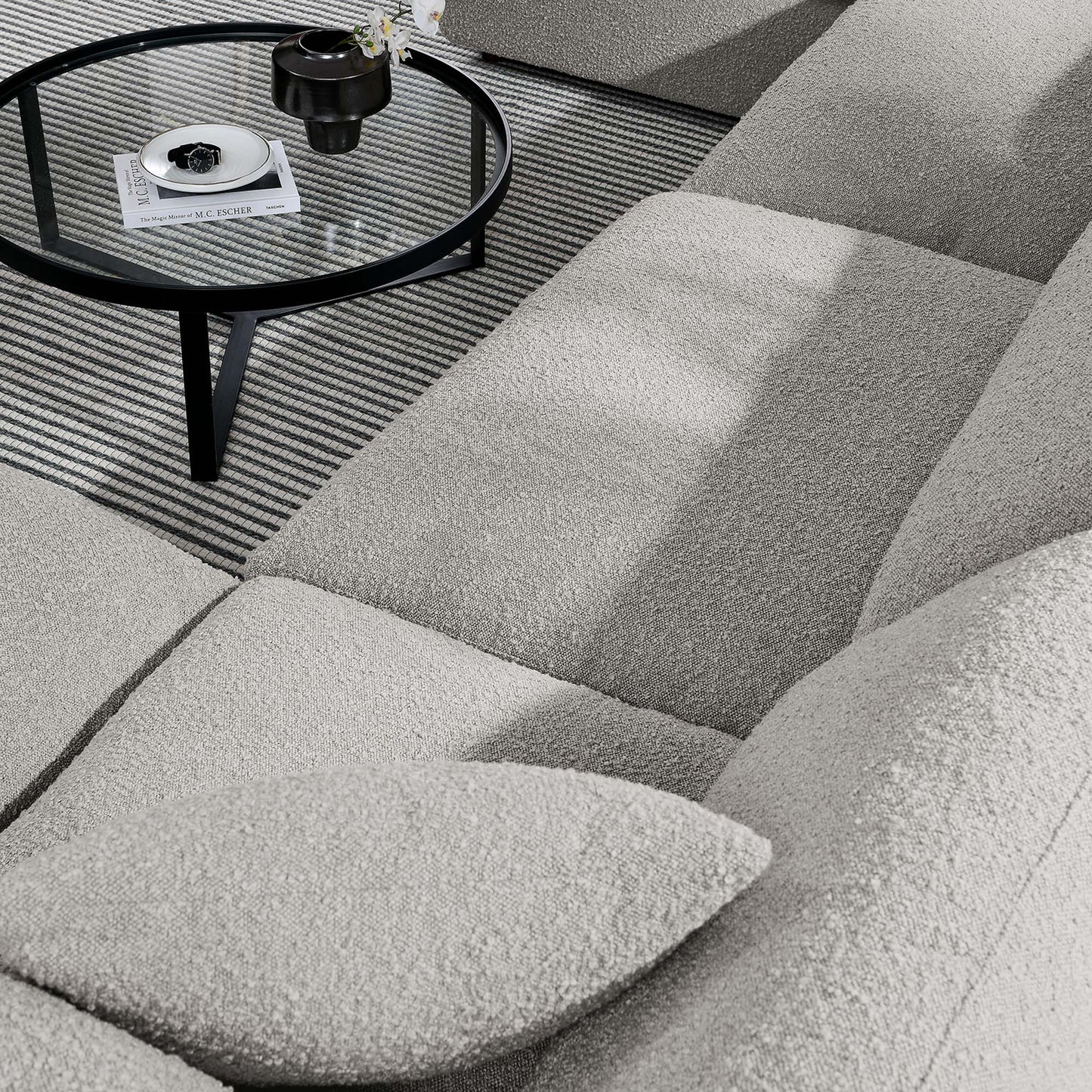 Light gray boucle sectional sofa with cushions, round glass coffee table on black metal legs, striped rug, magazine on table.