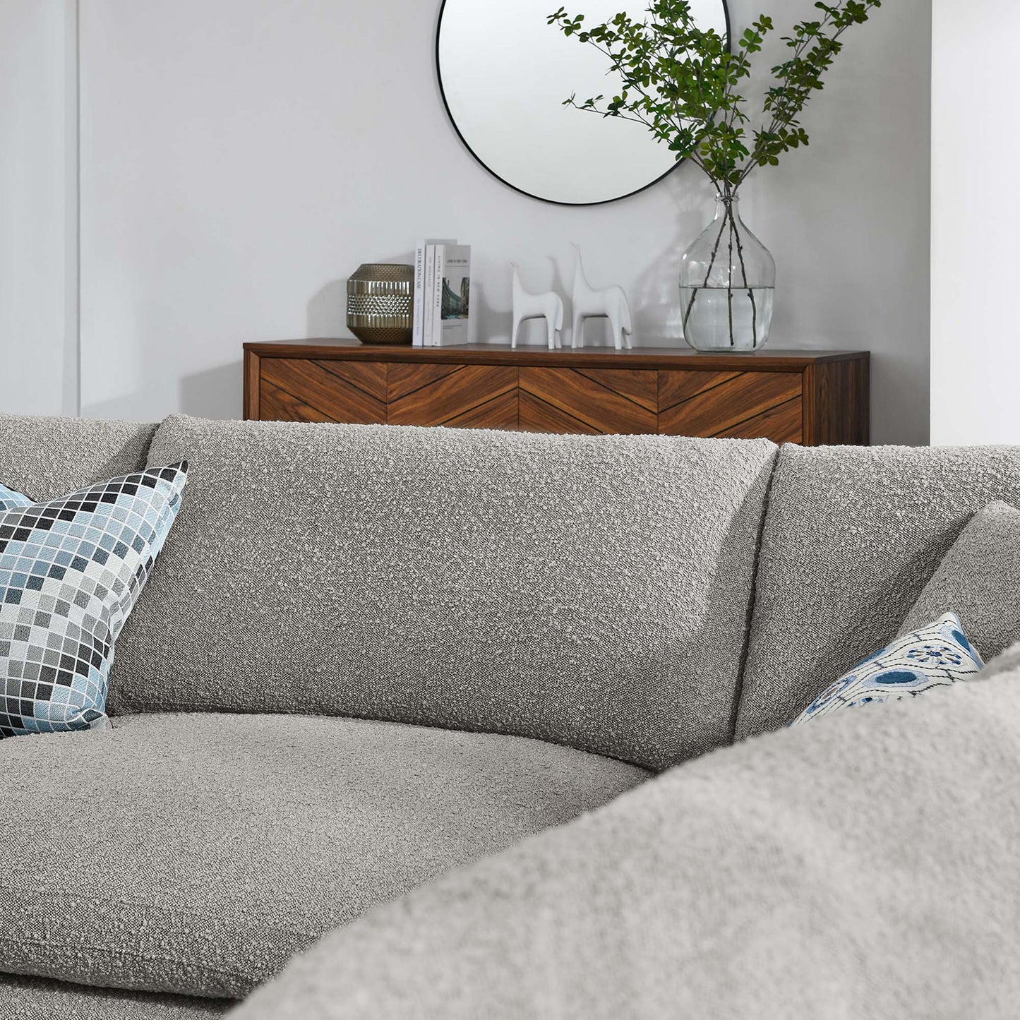 Textured light gray sectional sofa with blue geometric pillow, wooden console, round mirror, and glass vase with greenery.