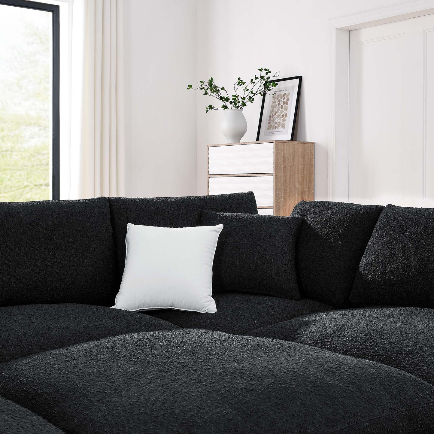Charcoal black fabric L-shaped sectional sofa with a white pillow in a bright modern living room light wood dresser and vase.