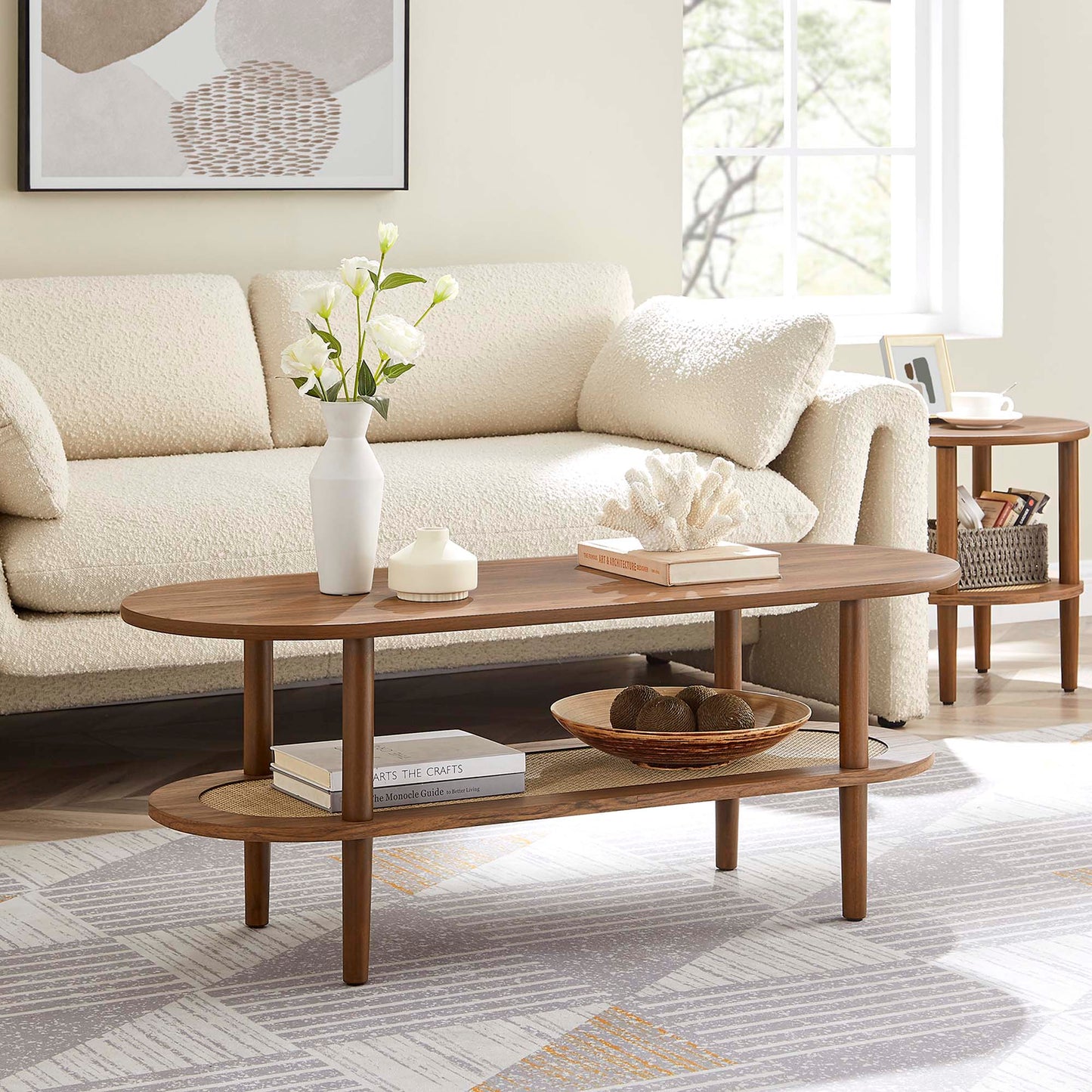 Walnut oval two-tier coffee table with lower shelf in beige living room; vase of white flowers, coral, books.