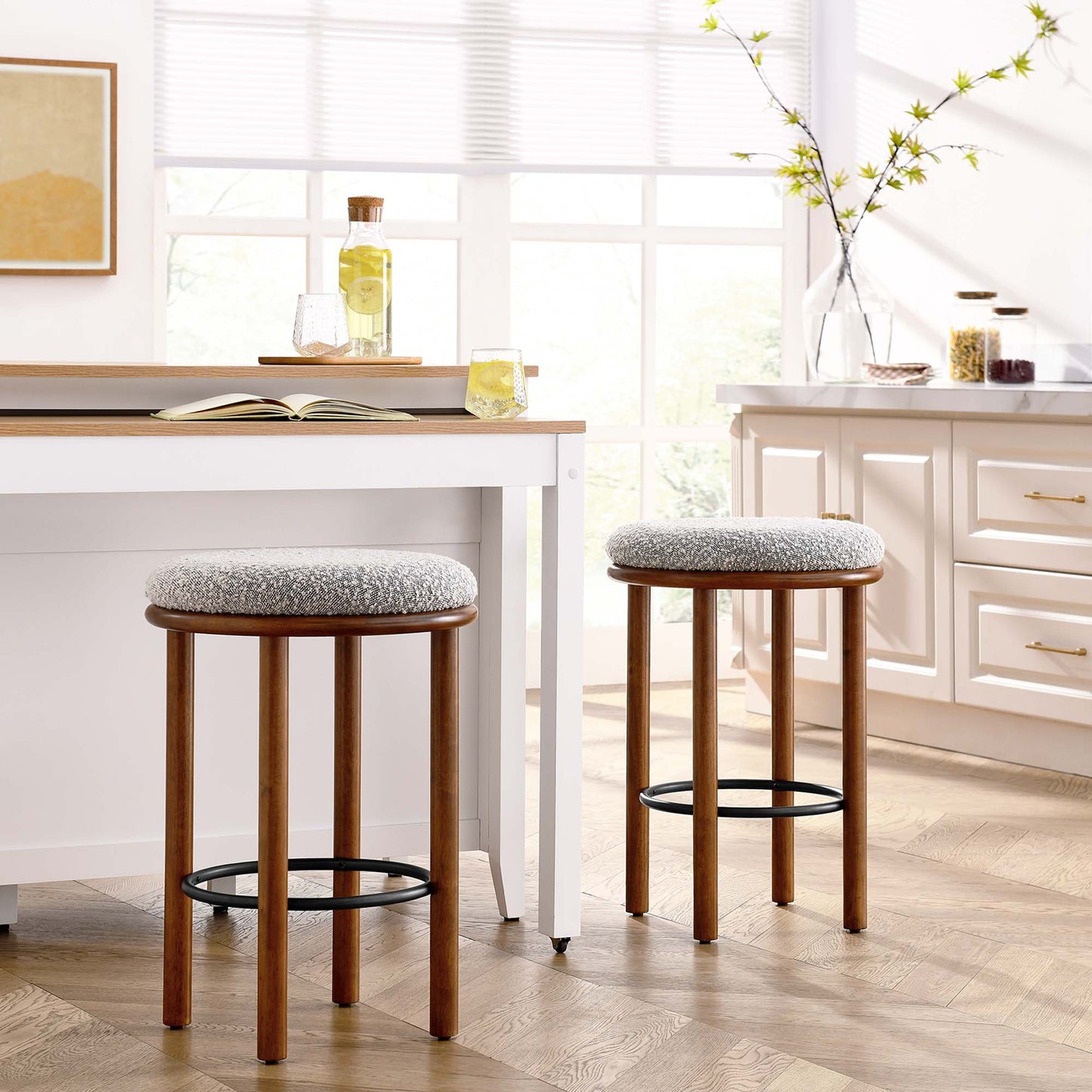 Pair of walnut wood counter stools with taupe upholstered seats and black circular footrests in bright white kitchen.