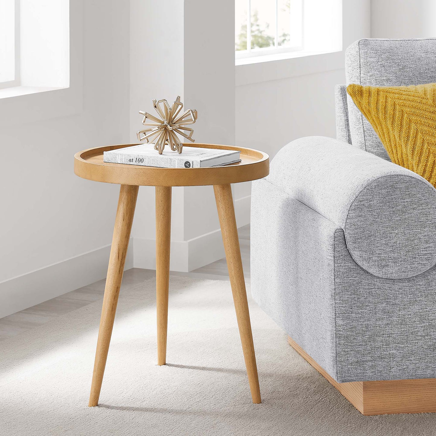 Natural wood round side table with three tapered legs, magazines on top, in a bright living room beside a gray sofa.