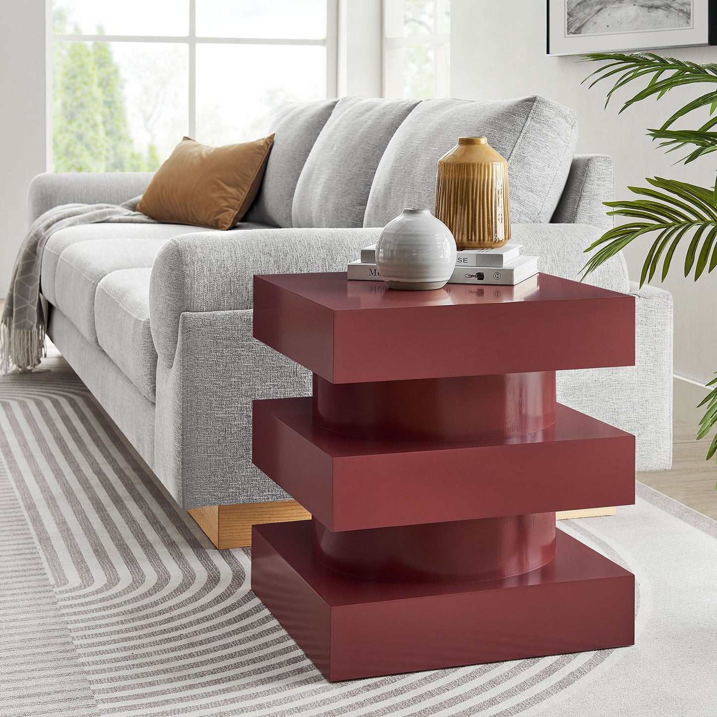 Glossy burgundy stacked end table with three wide platforms beside a light gray sofa; decor books and ceramic vases on top.