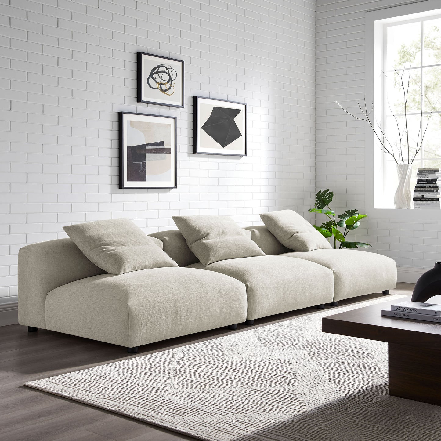 Beige linen three-seat sofa with plush cushions in a modern living room, abstract art, gray rug, dark wood coffee table.