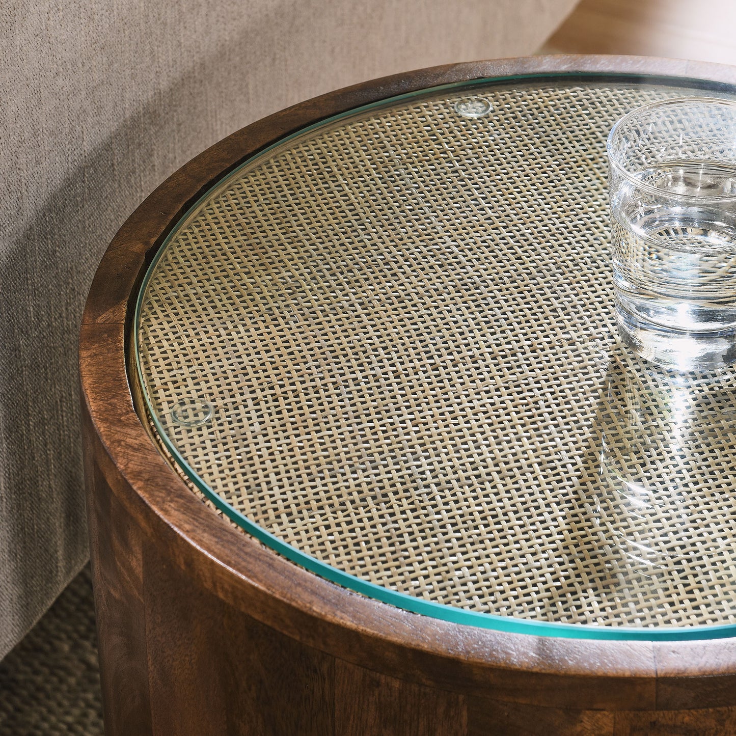 Round walnut wood rattan side table with a glass inset over a woven cane top; a glass of water rests near the edge.