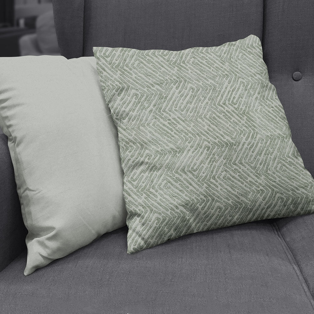 Two square throw pillows on a dark gray sofa, made with vapor velvet geometric fabric: left solid pale beige, right sage green with a chevron geometric pattern.
