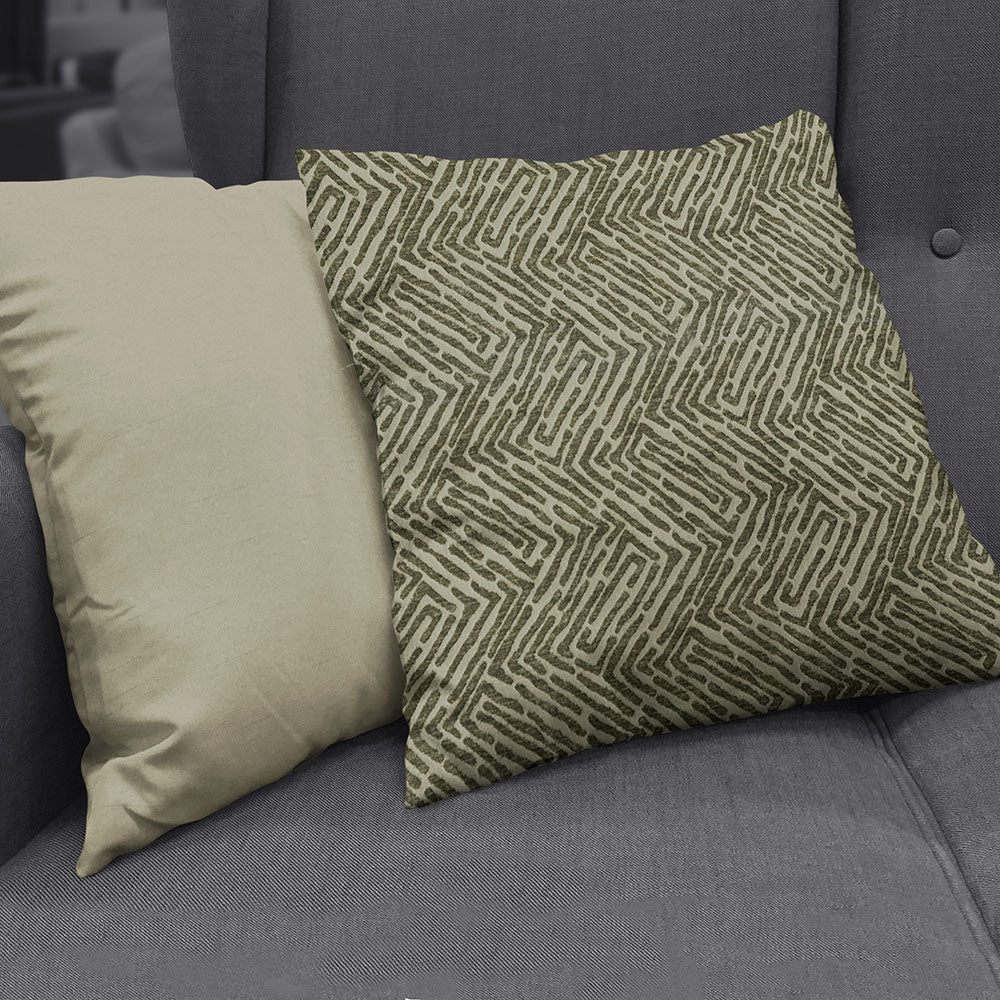 Olive geometric patterned square throw pillow beside a plain beige pillow on a gray sofa.