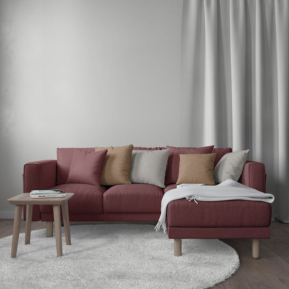 Maroon L-shaped sectional sofa with chaise, multicolor cushions, white throw, light wood legs, round white rug, side table, curtains.