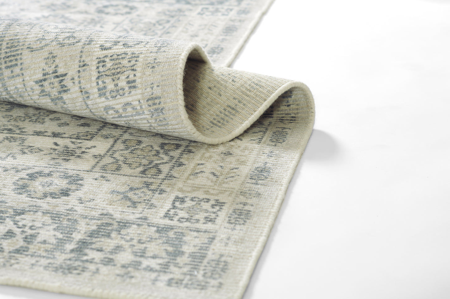 Gray and beige woven rug with faded geometric motifs, rolled edge showing texture.