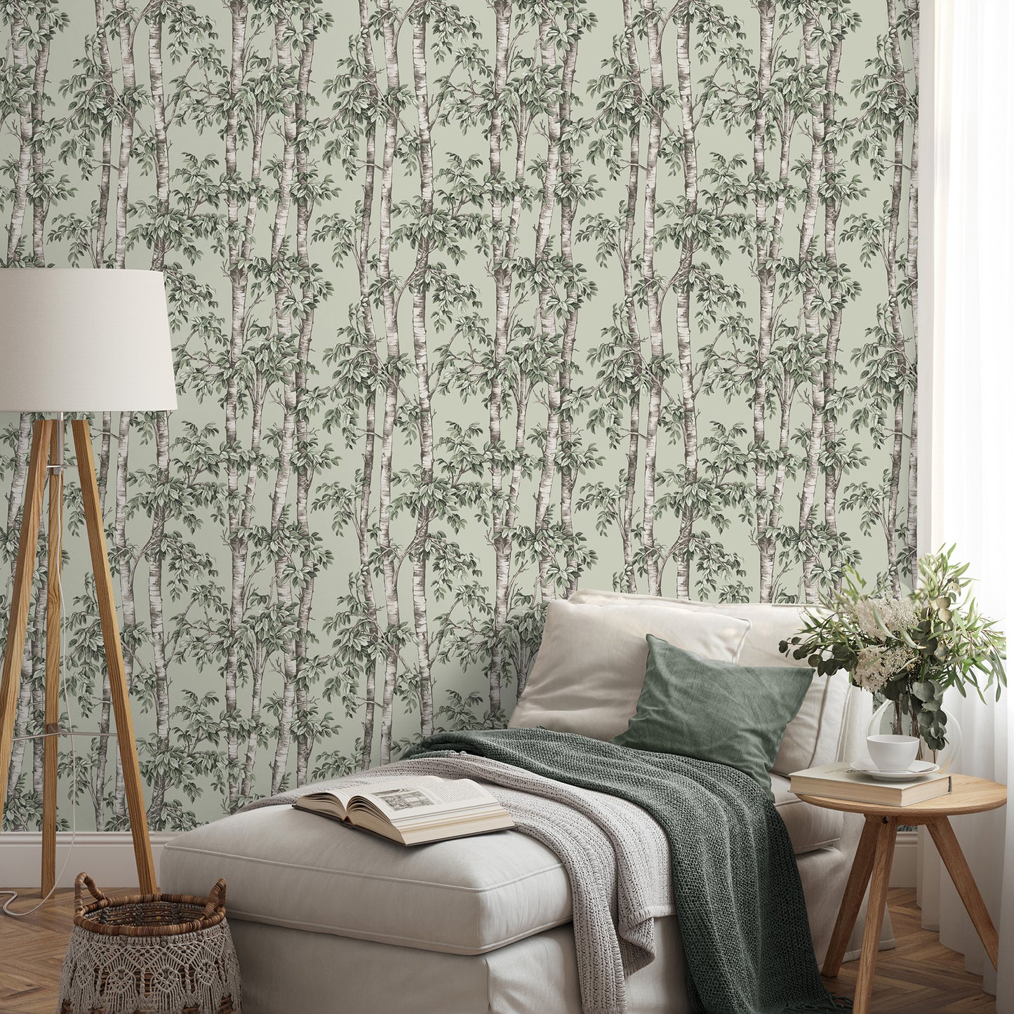 Beige chaise with green cushions and throws in a room featuring green trees botanical wallpaper, a wooden tripod floor lamp, and a round wood side table with a vase and a book.
