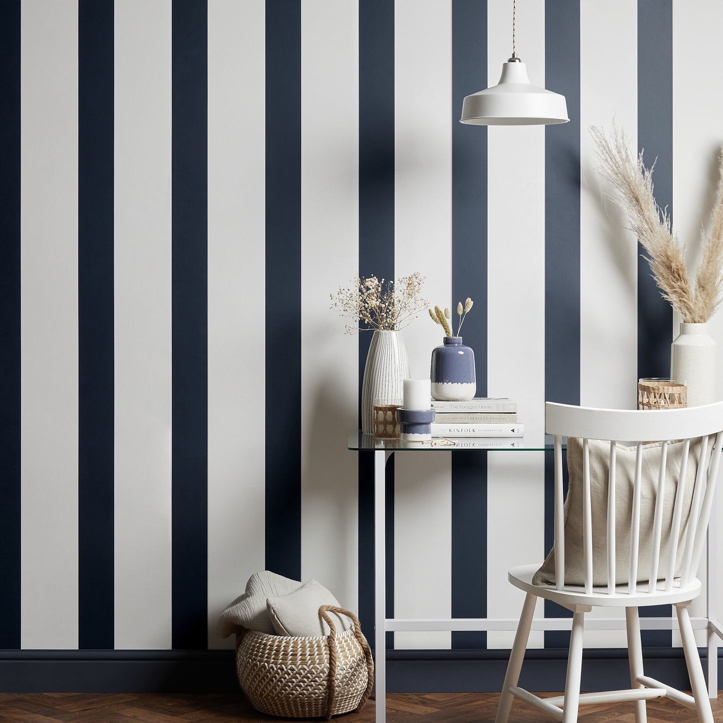 Blue and white vertical striped wallpaper with a glass table, white chair, ceramic vases, books, and a woven basket.