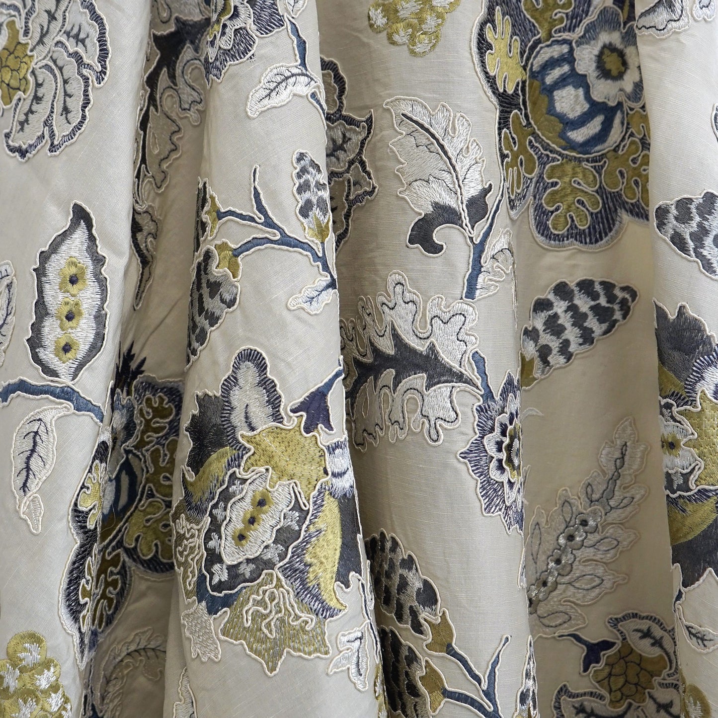 Beige fabric with dense metallic green floral embroidery; ornamental botanical pattern; silk texture.