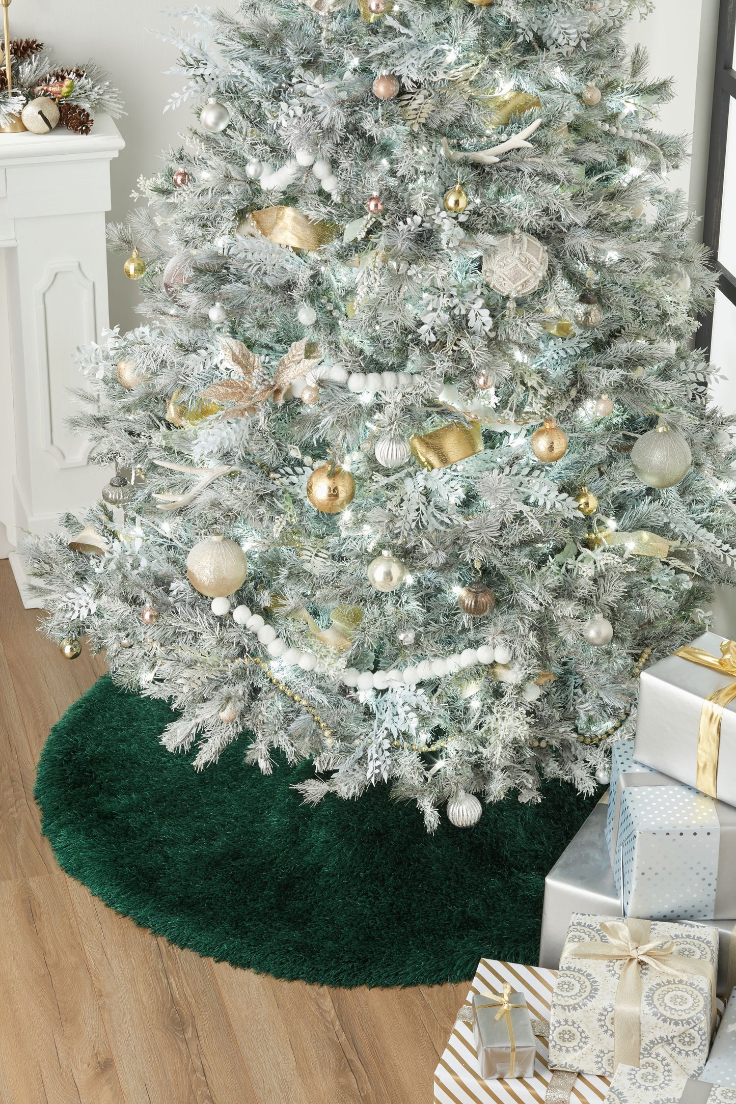 Snow-flocked artificial Christmas tree adorned with gold and white ornaments, placed on a round rug with wrapped gifts.