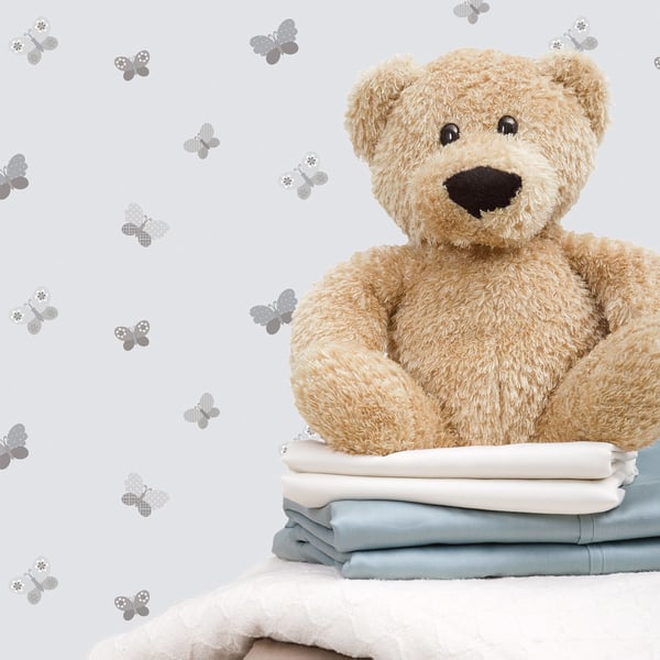 Soft beige teddy bear sits atop folded white and blue blankets, with a pale butterfly wallpaper background.