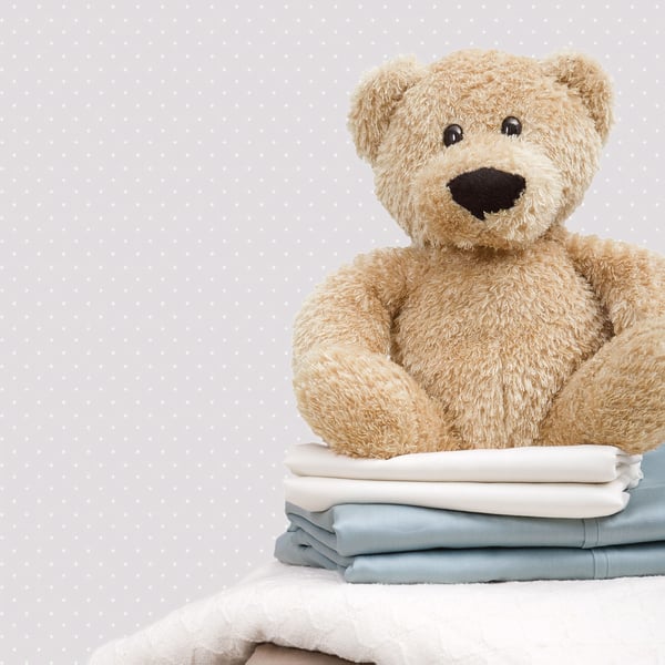 Beige plush teddy bear sits atop folded white and light-blue blankets against a pale polka-dot background.