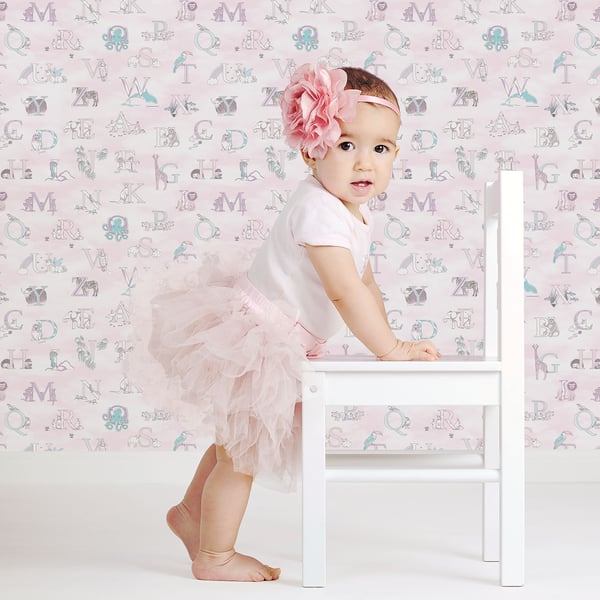 Baby girl in white top and pink tutu with a flower headband, standing beside a white wooden chair against pastel wallpaper.
